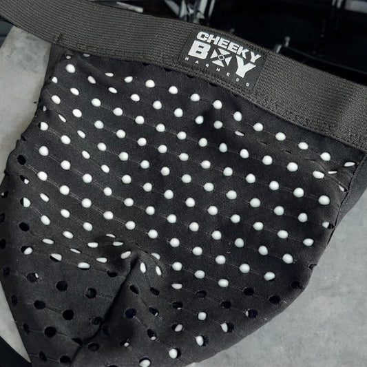 Man wearing a custom-fit black elastic body harness with matching bicep bands, featuring metal rings details and diagonal strap design. Styled with black mesh jockstrap underwear. Handmade by Cheeky Boy Harness