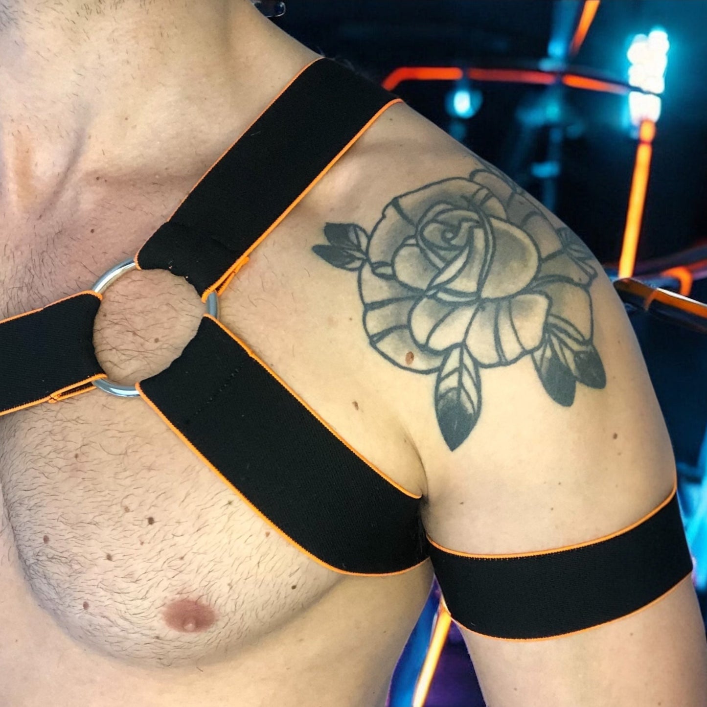 Man wearing a handmade black chest harness with glowing neon orange edges, styled in a Spartan warrior design. Includes matching bicep bands. Photographed in a neon-lit club setting. The harness is made to measure and glows under UV light, ideal for clubbing and festival wear.