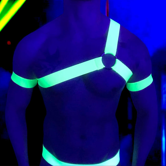 A man wearing a neon yellow harness and matching accessories under blacklight, causing the outfit to glow vibrantly. The outfit includes a chest harness with a central ring, armbands, thigh bands, and a waistband on black briefs. The background is a dark club or rave setting with purple and yellow lighting effects