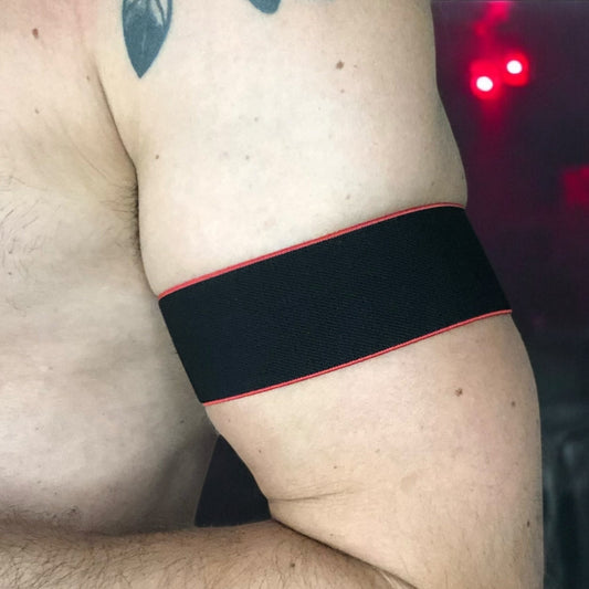 Man wearing a handmade black chest harness with glowing neon orange edges, styled in a Spartan warrior design. Includes matching bicep bands. Photographed in a neon-lit club setting. The harness is made to measure and glows under UV light, ideal for clubbing and festival wear.