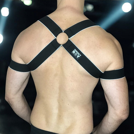 Man wearing a handmade black elastic chest harness with white trim and matching bicep bands by Cheeky Boy Harness, modeled in a dark clubwear setting.