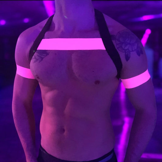 A man wearing a handmade, made-to-measure black and neon pink elastic chest harness with matching jockstrap and bicep bands by Cheeky Boy Harness, shown in a dark club setting with glowing pink UV lights
