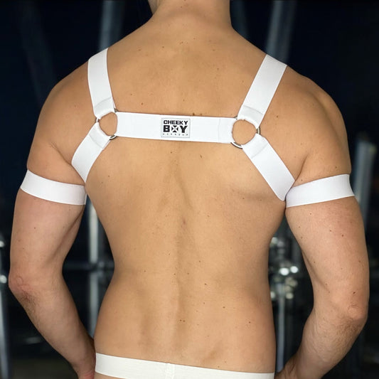 Man wearing a white elastic chest harness with 6 metal rings throughout with matching bicep bands and a white mesh jockstrap, posed in a nightclub setting, by Cheeky Boy Harness.
