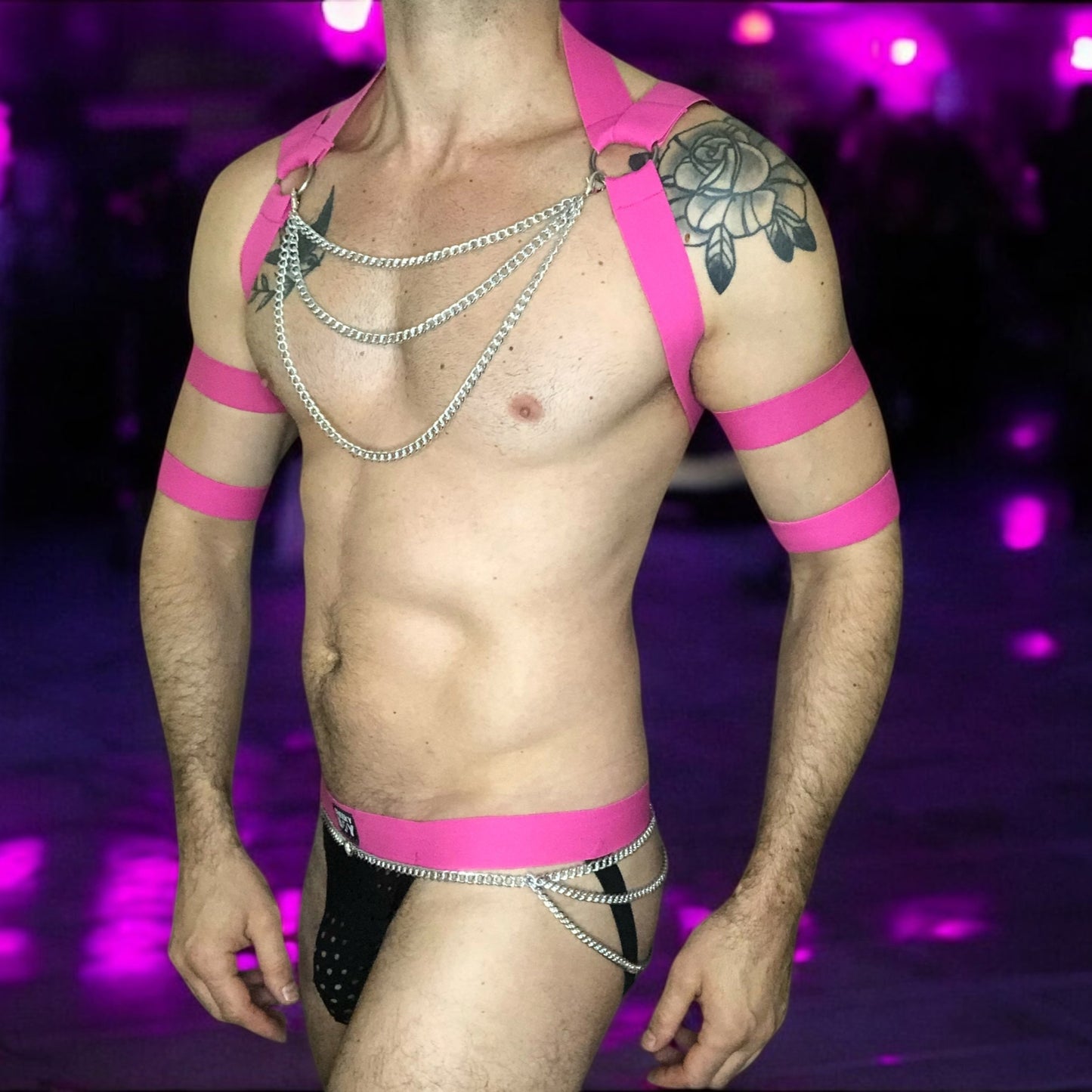 Model wearing a hot pink elastic chest harness with silver chains on chest and waistband, matching bicep bands, and a black mesh jockstrap with pink waistband by Cheeky Boy Harness.