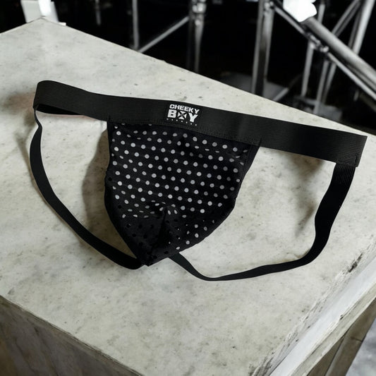 Man wearing a custom-fit black elastic body harness with matching bicep bands, featuring metal rings details and diagonal strap design. Styled with black mesh jockstrap underwear. Handmade by Cheeky Boy Harness