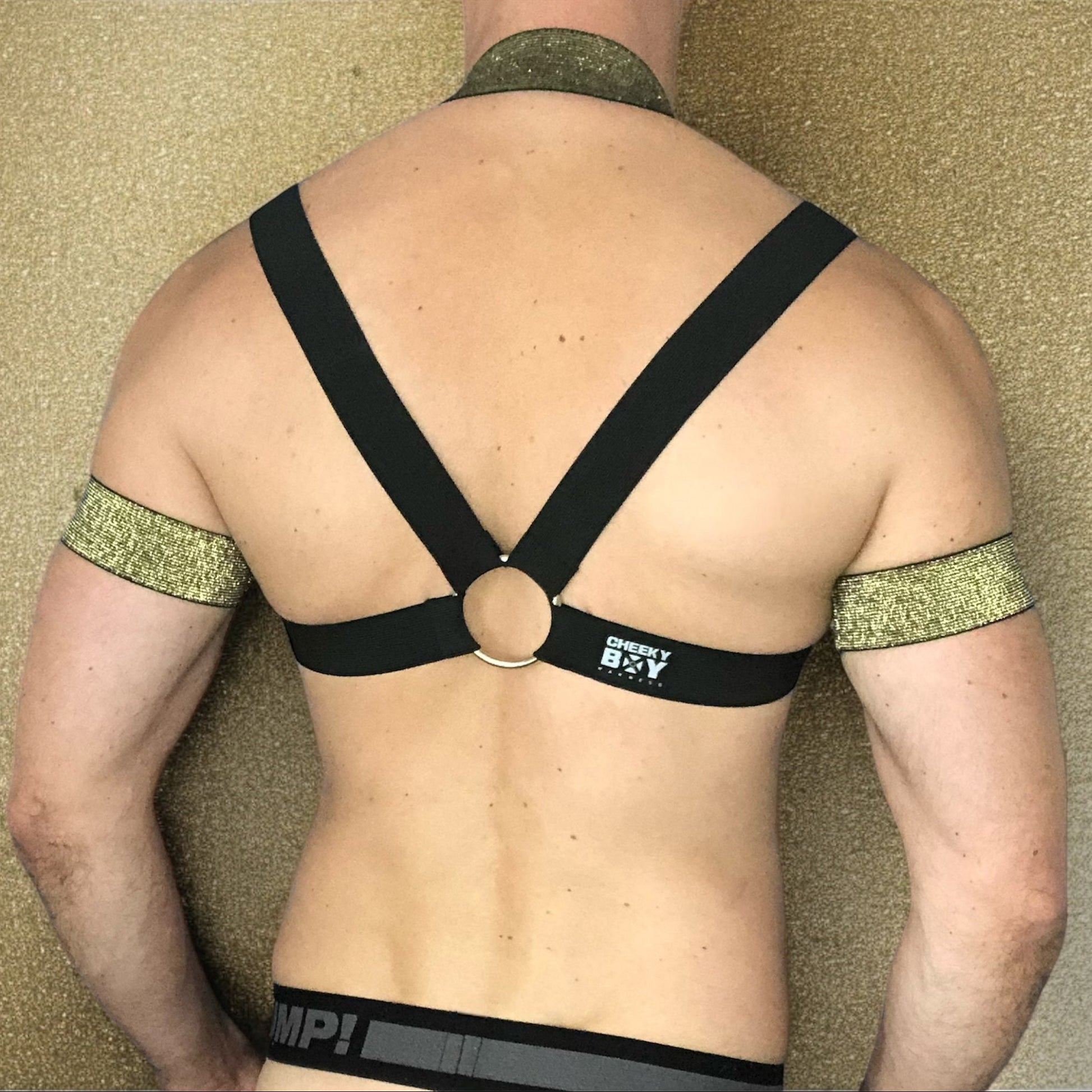 Model wearing a custom-made black & gold elastic body harness with metal rings and matching jockstrap by Cheeky Boy Harness. Includes gold bicep bands and leg straps, designed for clubwear, fetish events, and festivals