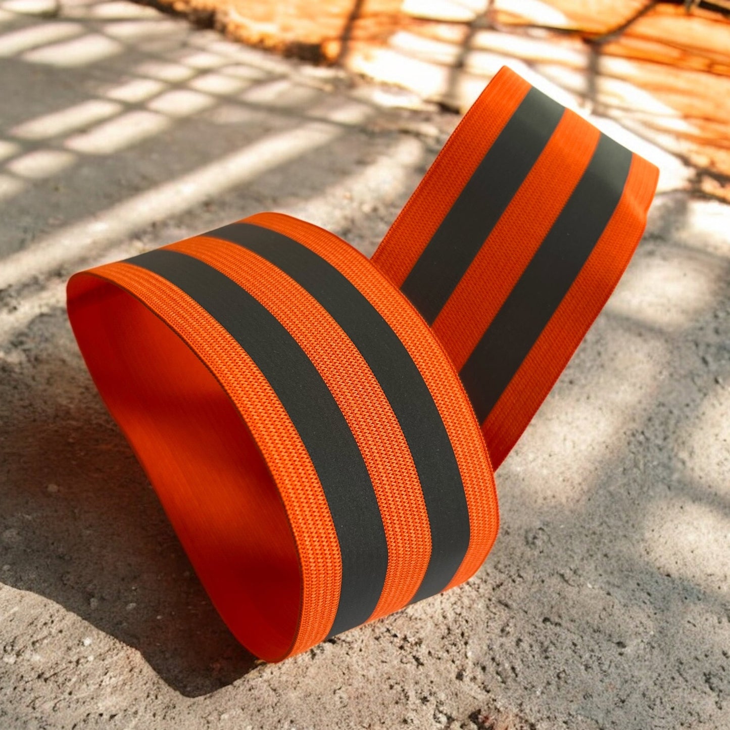 Bright orange Hi Vis bicep bands with reflective stripes, styled as a loop and displayed on a sunlit concrete surface, suitable for use as bicep or thigh bands.