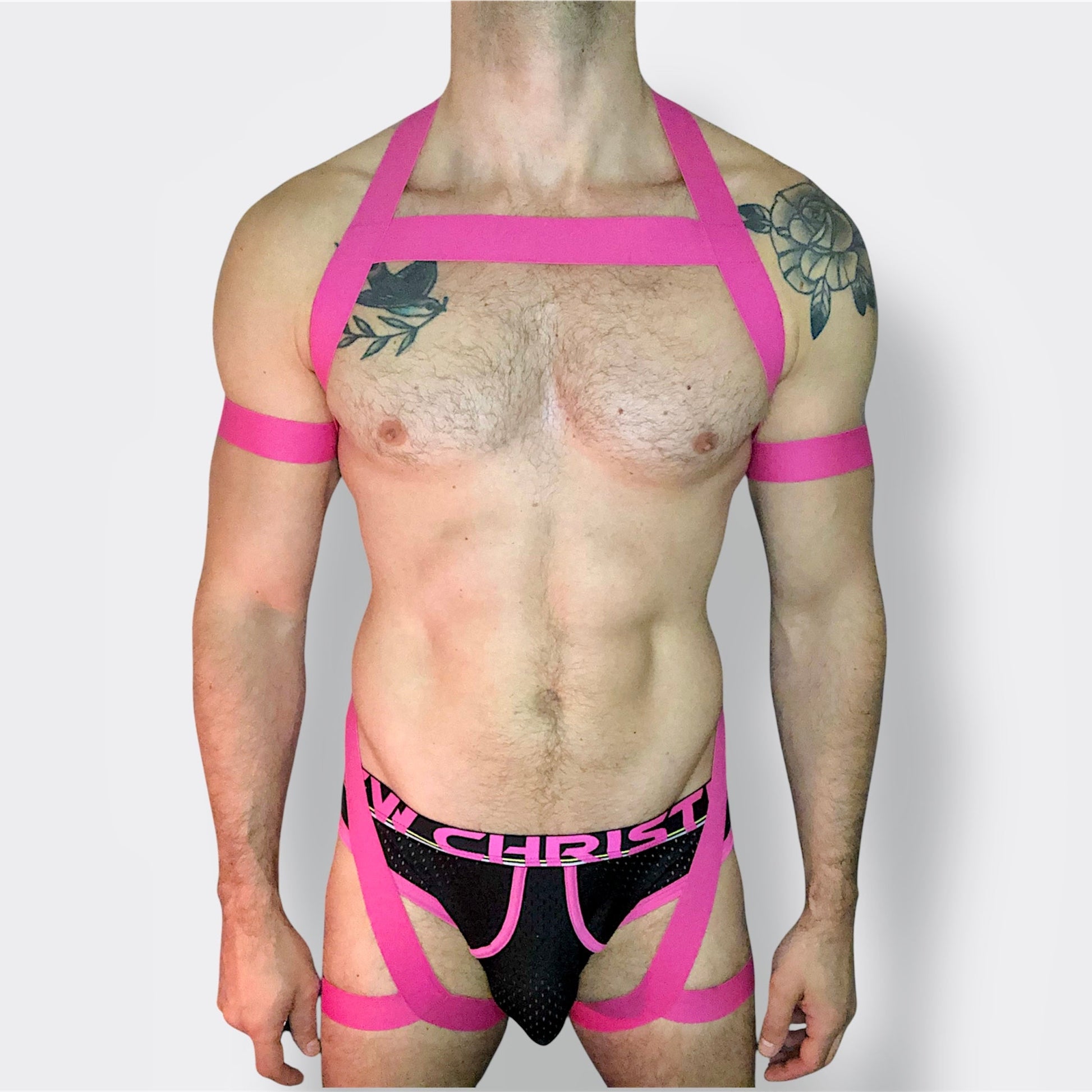 Model wearing a custom hot pink elastic body harness by Cheeky Boy Harness, featuring a silver chest ring, matching bicep bands, and thigh straps, styled with a pink mesh jockstrap.