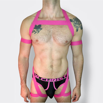 Model wearing a custom hot pink elastic body harness by Cheeky Boy Harness, featuring a silver chest ring, matching bicep bands, and thigh straps, styled with a pink mesh jockstrap.