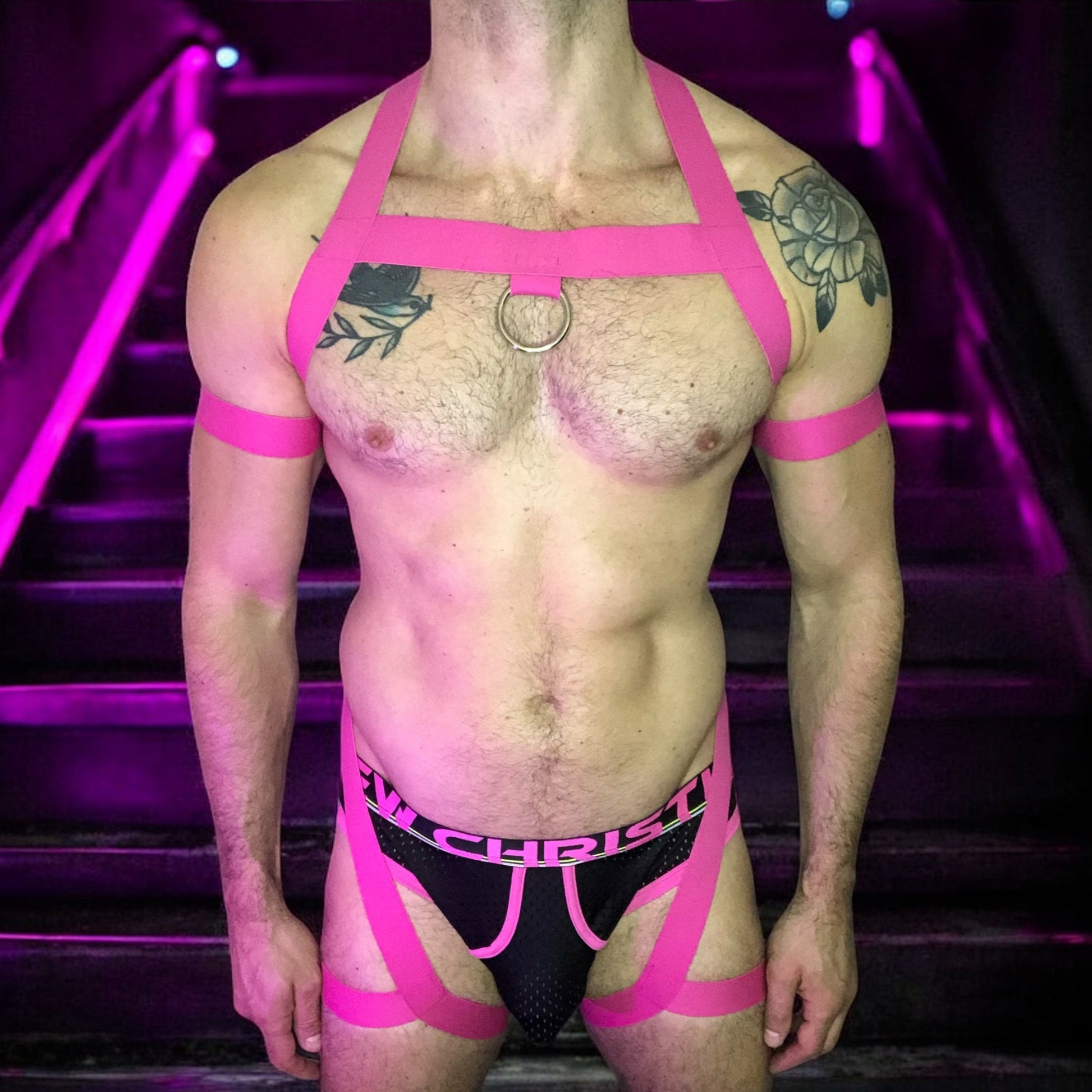 Model wearing a custom hot pink elastic body harness by Cheeky Boy Harness, featuring a silver chest ring, matching bicep bands, and thigh straps, styled with a pink mesh jockstrap.