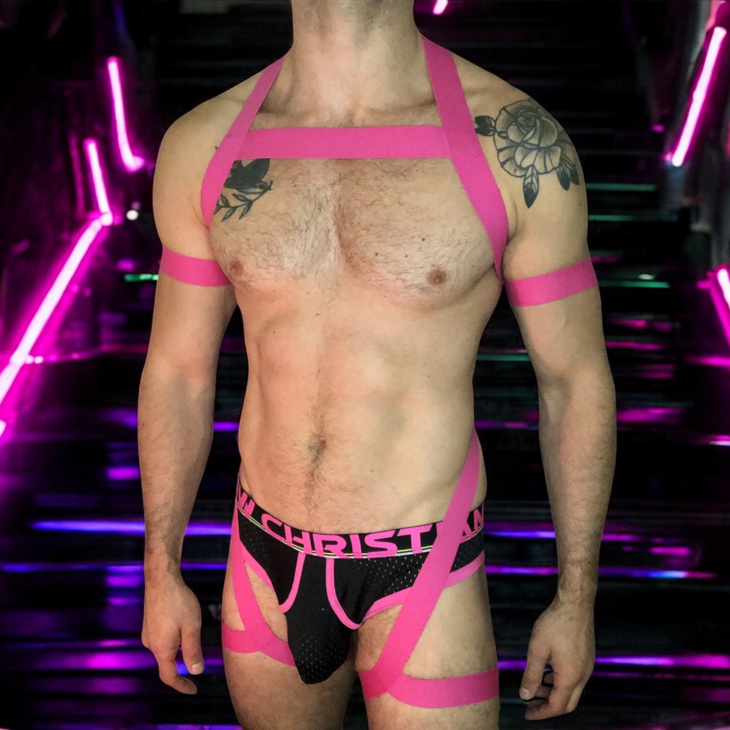 Model wearing a custom hot pink elastic body harness by Cheeky Boy Harness, featuring a silver chest ring, matching bicep bands, and thigh straps, styled with a pink mesh jockstrap.