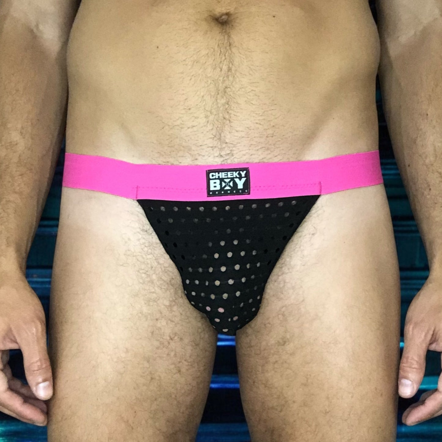 Model wearing a custom hot pink elastic body harness by Cheeky Boy Harness, featuring a silver chest ring, matching bicep bands, and thigh straps, styled with a pink mesh jockstrap.