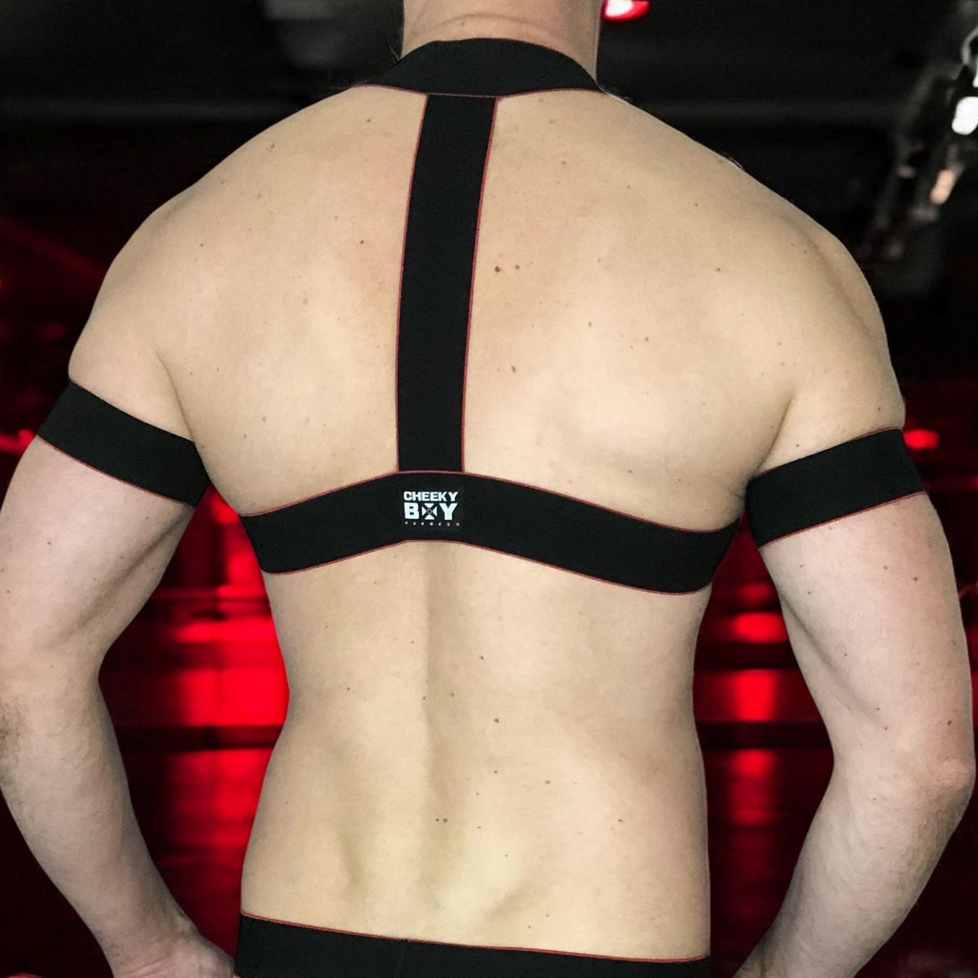 Toned model Wearing Made-to-Measure Black and Red Elastic Chest Harness with Metal Chest Ring by Cheeky Boy Harness, Styled for Fetish Clubwear in Red-Lit Venue