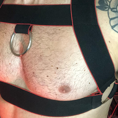 Toned model Wearing Made-to-Measure Black and Red Elastic Chest Harness with Metal Chest Ring by Cheeky Boy Harness, Styled for Fetish Clubwear in Red-Lit Venue