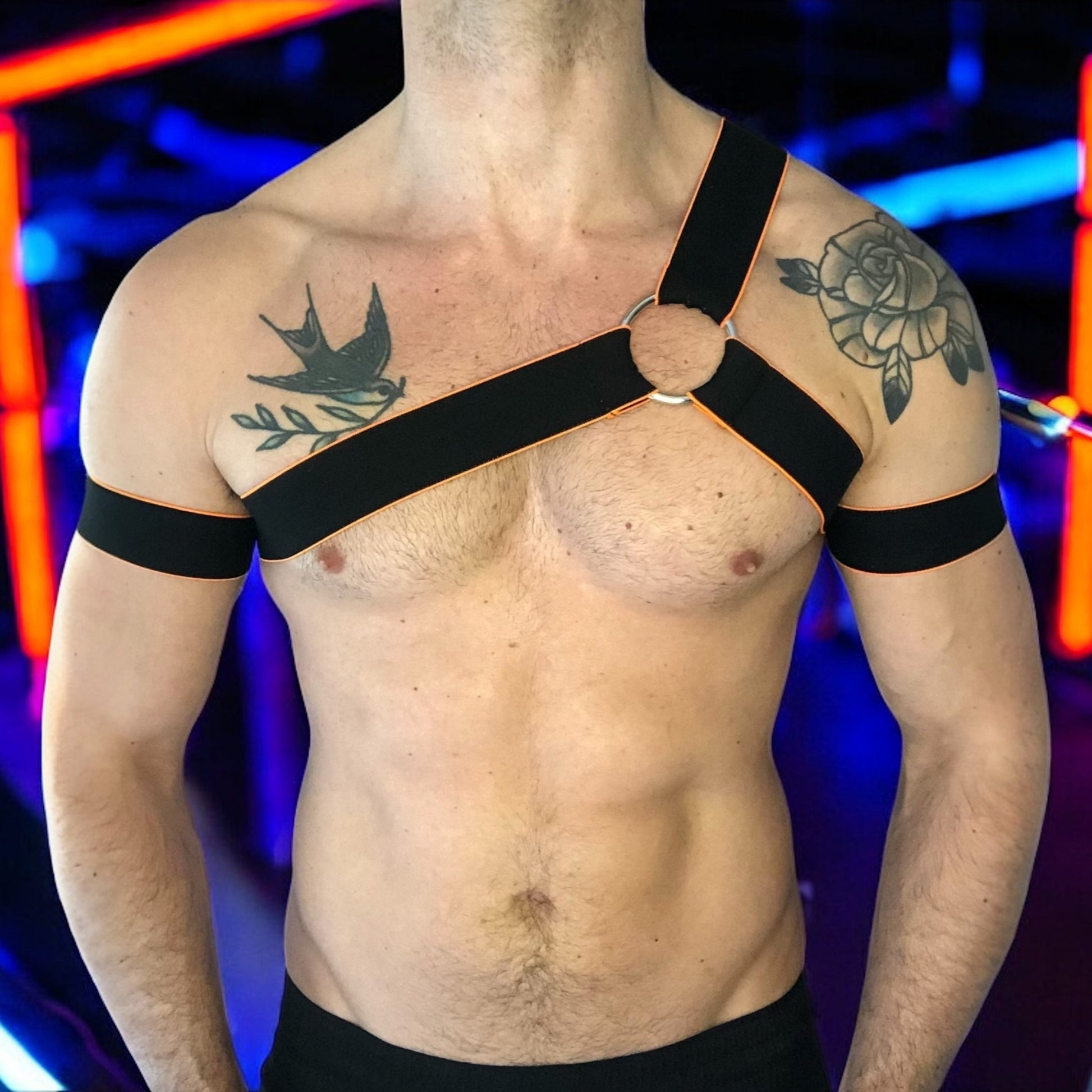 Man wearing a handmade black chest harness with glowing neon orange edges, styled in a Spartan warrior design. Includes matching bicep bands. Photographed in a neon-lit club setting. The harness is made to measure and glows under UV light, ideal for clubbing and festival wear.