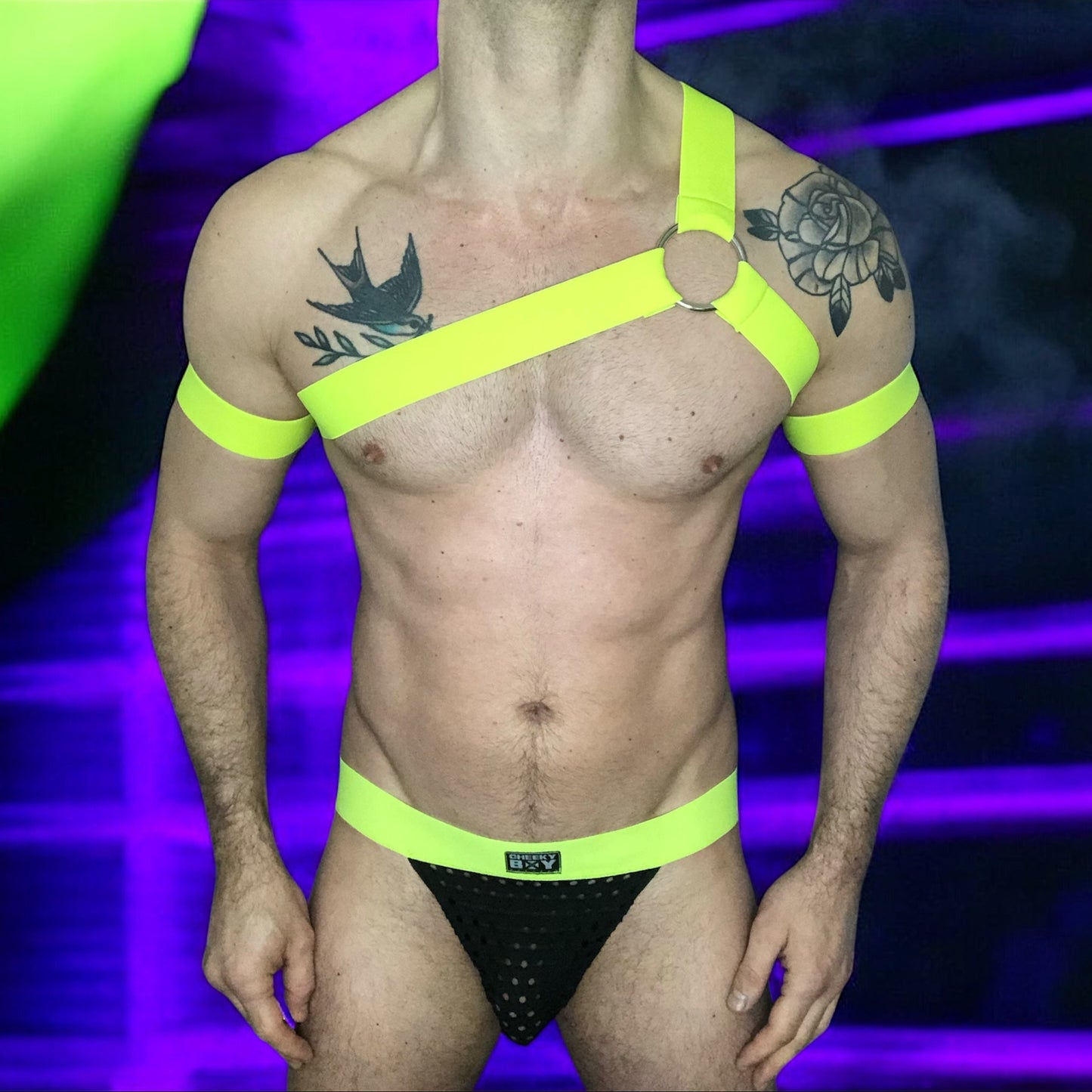 A man wearing a neon yellow harness and matching accessories under blacklight, causing the outfit to glow vibrantly. The outfit includes a chest harness with a central ring, armbands, thigh bands, and a waistband on black briefs. The background is a dark club or rave setting with purple and yellow lighting effects