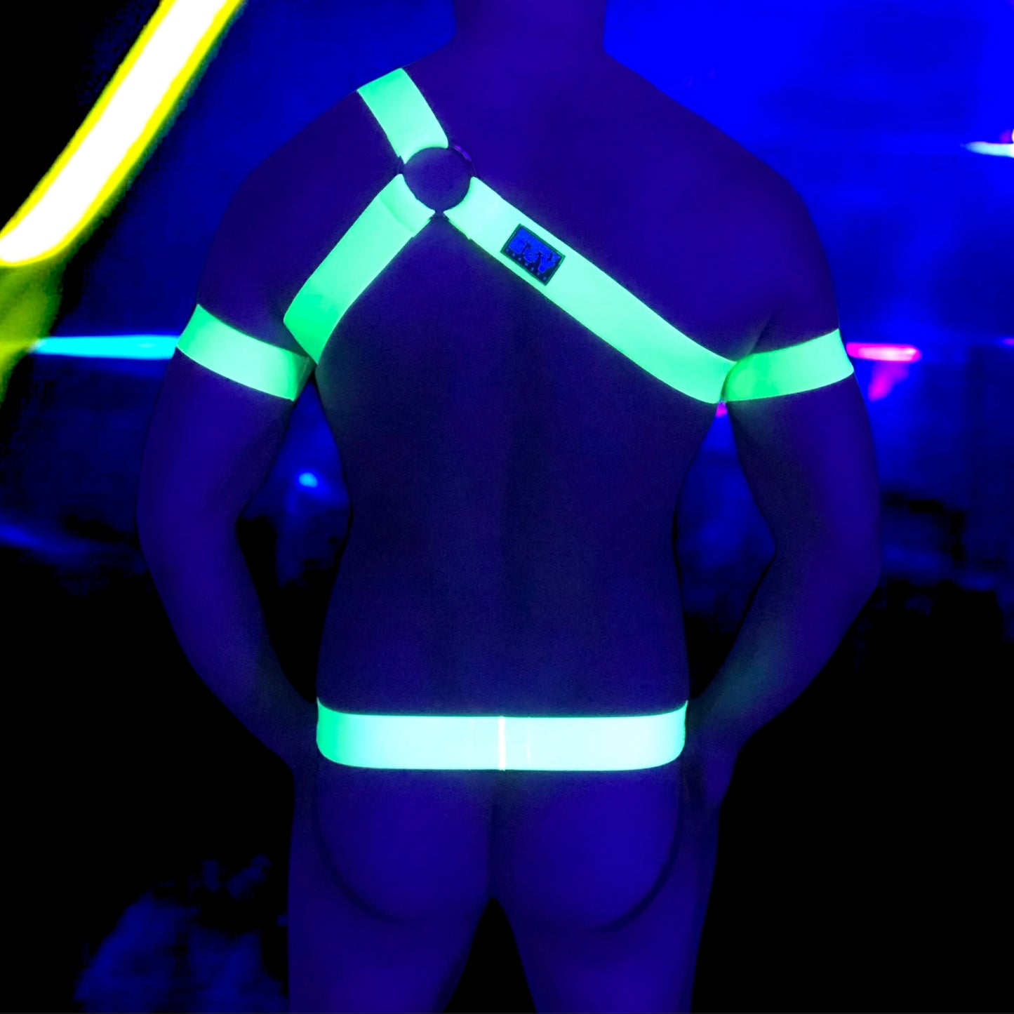 A man wearing a neon yellow harness and matching accessories under blacklight, causing the outfit to glow vibrantly. The outfit includes a chest harness with a central ring, armbands, thigh bands, and a waistband on black briefs. The background is a dark club or rave setting with purple and yellow lighting effects