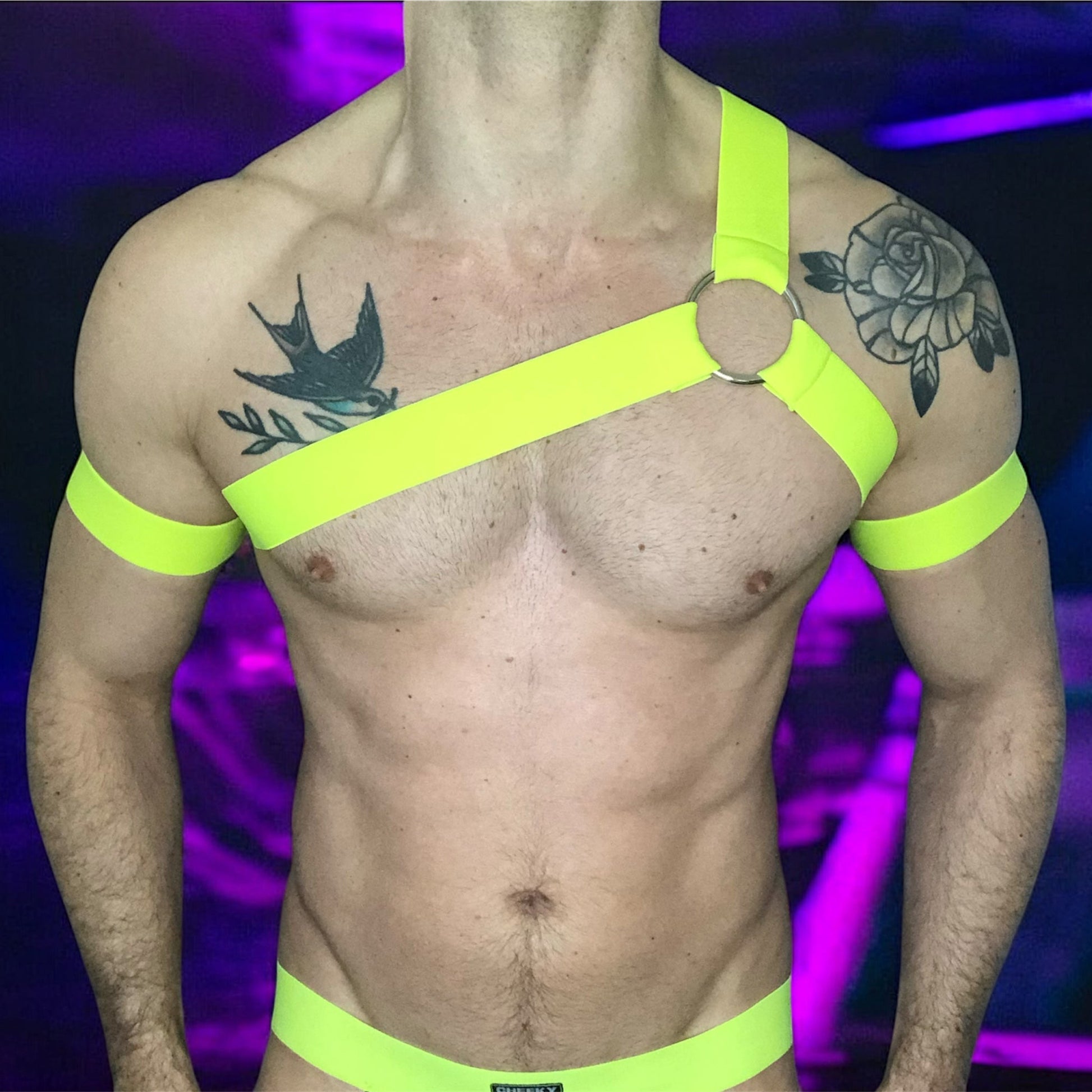 A man wearing a neon yellow harness and matching accessories under blacklight, causing the outfit to glow vibrantly. The outfit includes a chest harness with a central ring, armbands, thigh bands, and a waistband on black briefs. The background is a dark club or rave setting with purple and yellow lighting effects