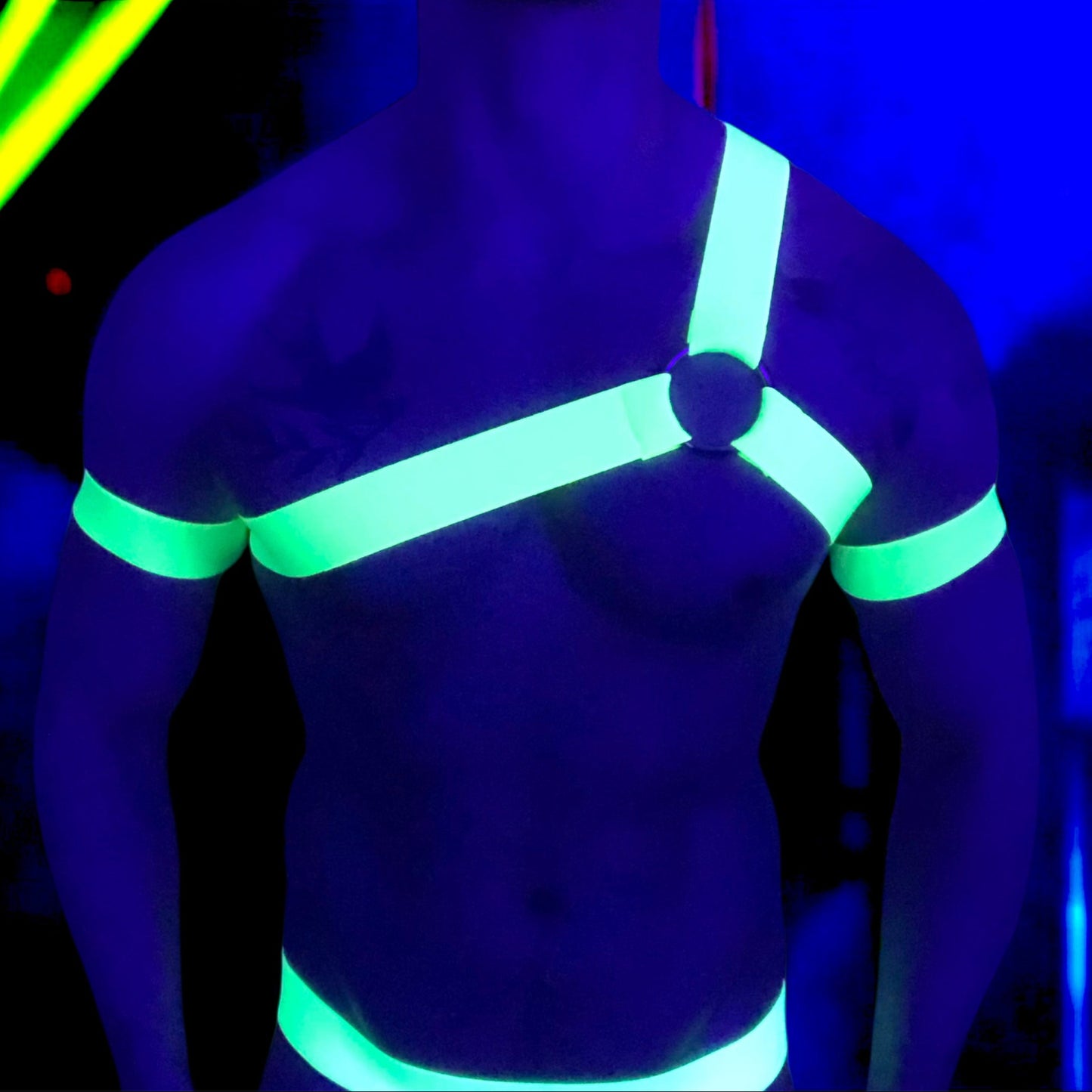 A man wearing a neon yellow harness and matching accessories under blacklight, causing the outfit to glow vibrantly. The outfit includes a chest harness with a central ring, armbands, thigh bands, and a waistband on black briefs. The background is a dark club or rave setting with purple and yellow lighting effects