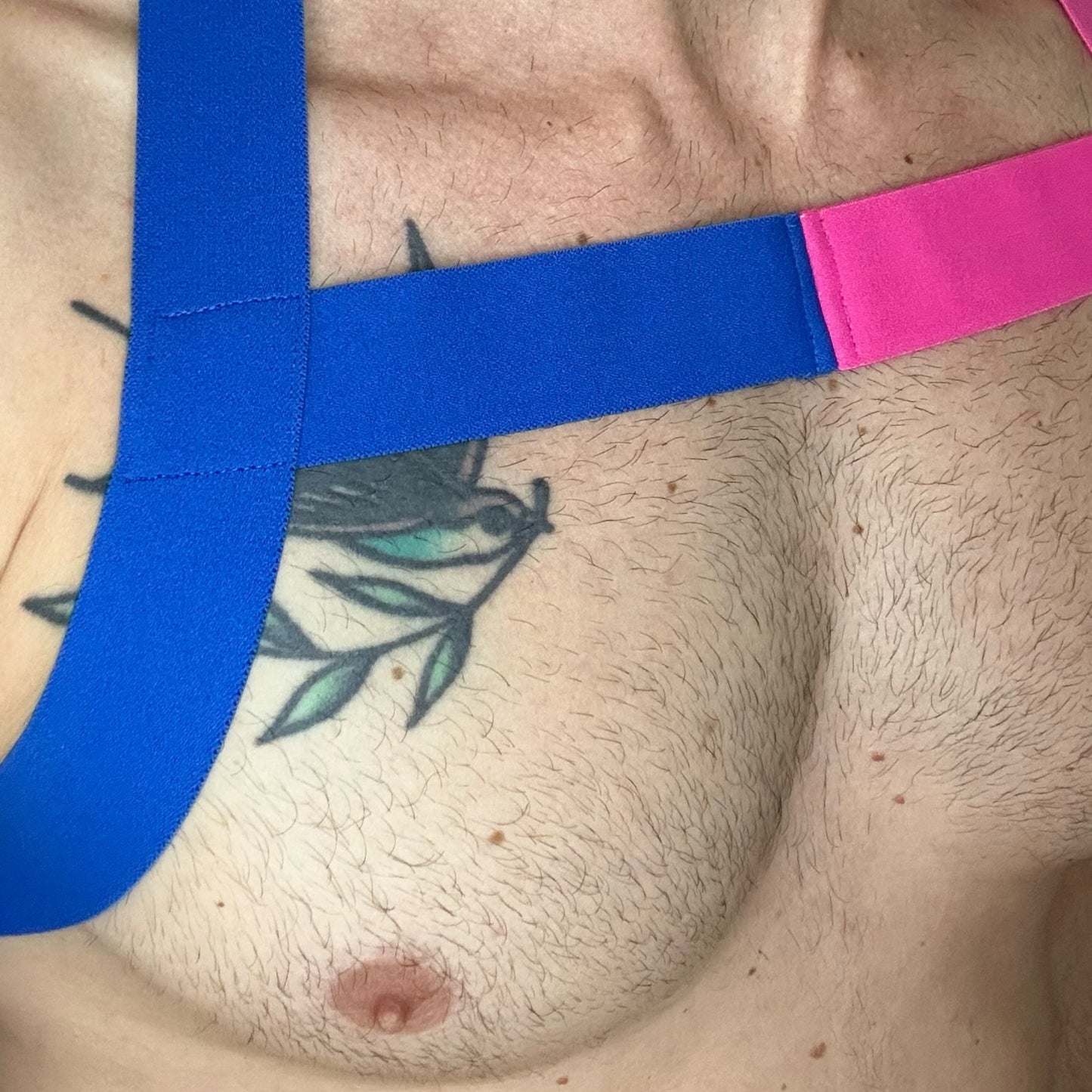 Model wearing a handmade two-tone half pink and half blue full body harness with matching bicep and thigh bands, paired with a white mesh jockstrap, posing in a neon-lit club setting
