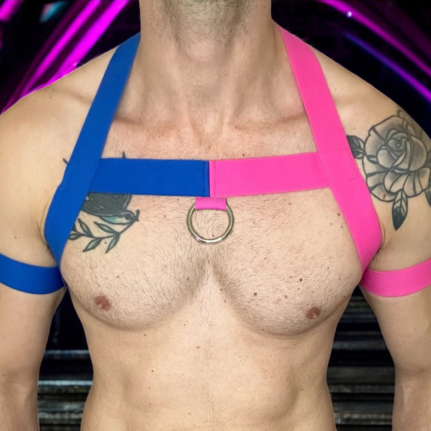 Model wearing a handmade two-tone half pink and half blue full body harness with matching bicep and thigh bands, paired with a white mesh jockstrap, posing in a neon-lit club setting