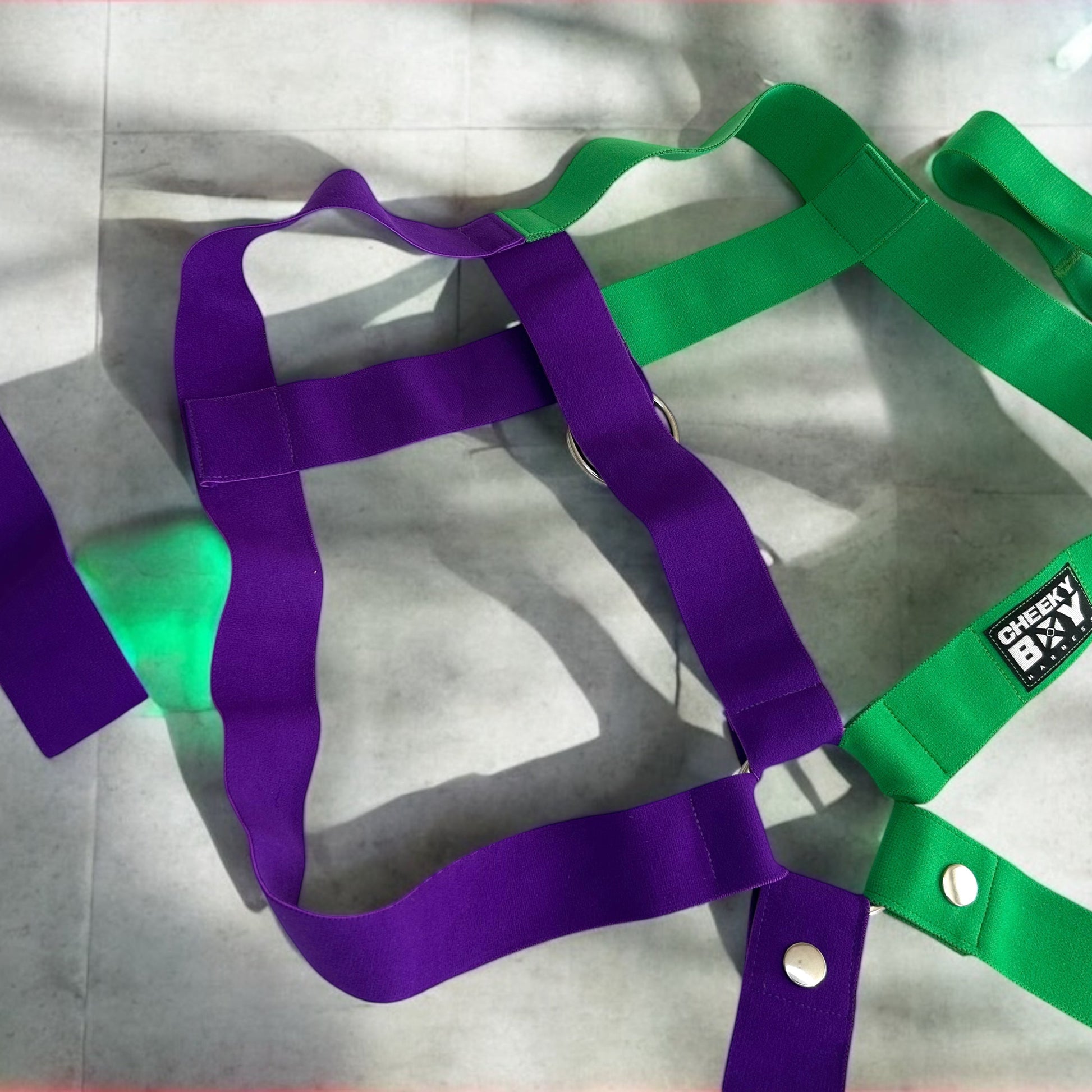 Model wearing a handmade two-tone half green and half purple 
 full body harness with matching bicep and thigh bands, paired with a white mesh jockstrap, posing in a neon-lit club setting