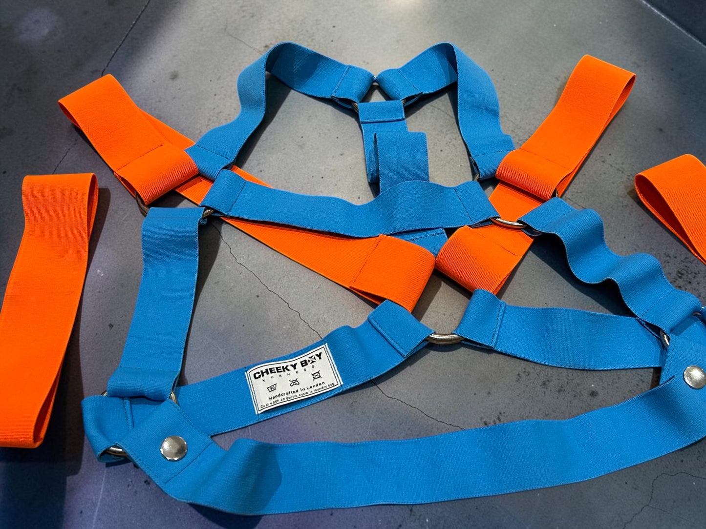 Flat lay of the turquoise men’s chest harness set with Neon Orange shoulder sections, matching bands, metal rings and removable lower strap.