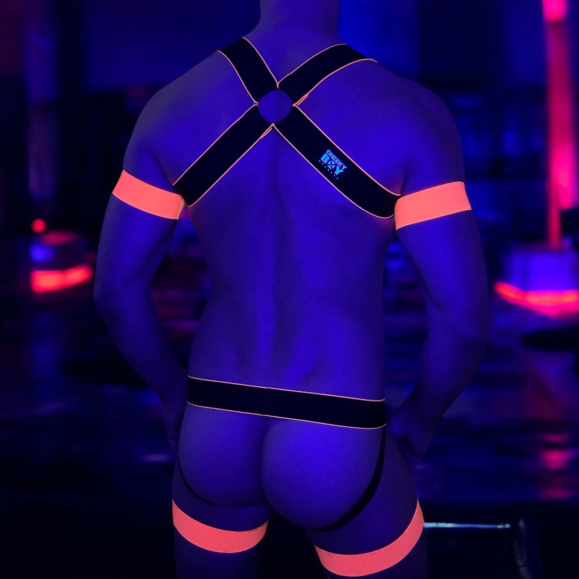 Man wearing a handmade black and neon orange elastic body harness with matching bicep bands, jockstrap, and thigh bands by Cheeky Boy Harness in a nightclub setting