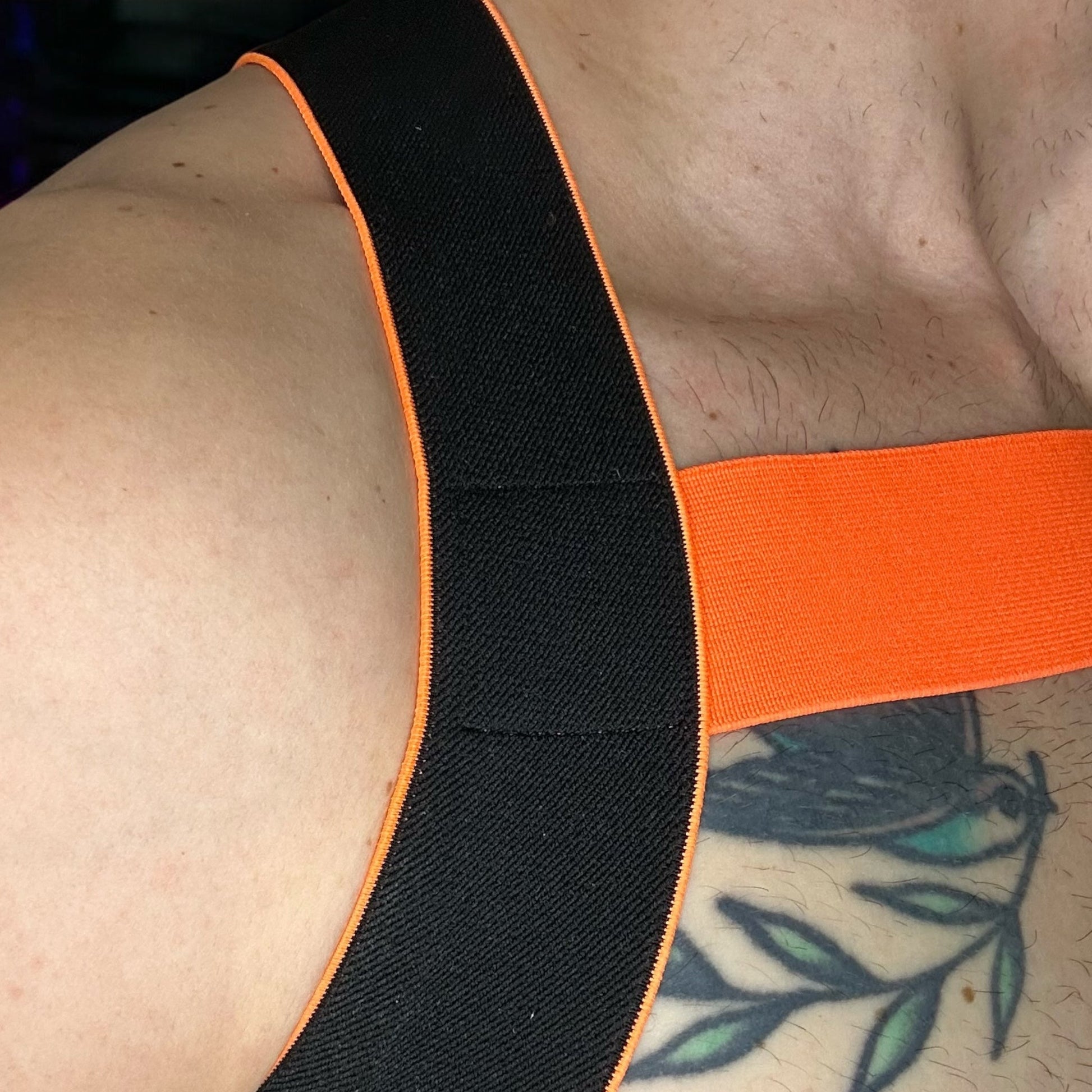 Man wearing a handmade black and neon orange elastic body harness with matching bicep bands, jockstrap, and thigh bands by Cheeky Boy Harness in a nightclub setting