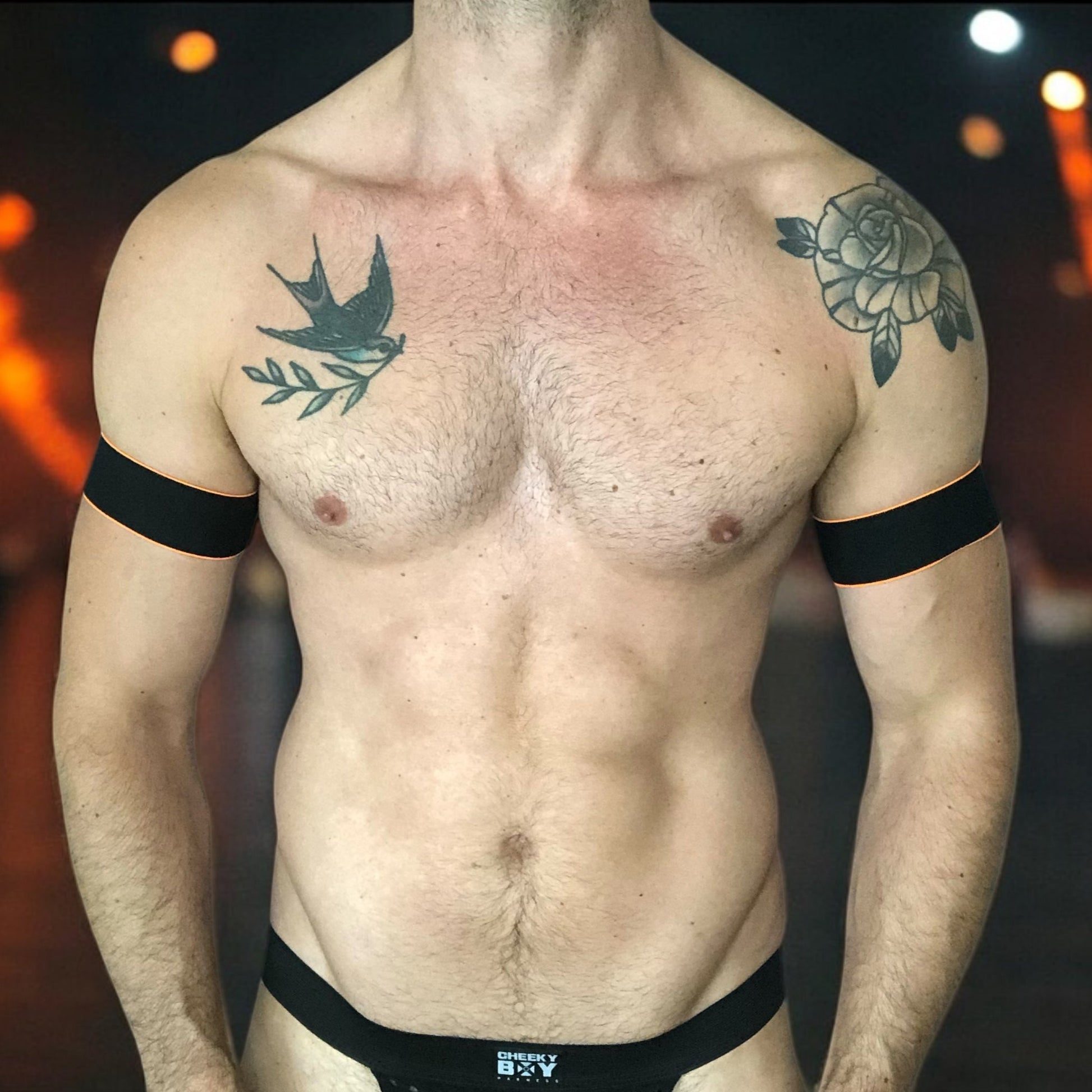 Man wearing a handmade black chest harness with glowing neon orange edges, styled in a Spartan warrior design. Includes matching bicep bands. Photographed in a neon-lit club setting. The harness is made to measure and glows under UV light, ideal for clubbing and festival wear.