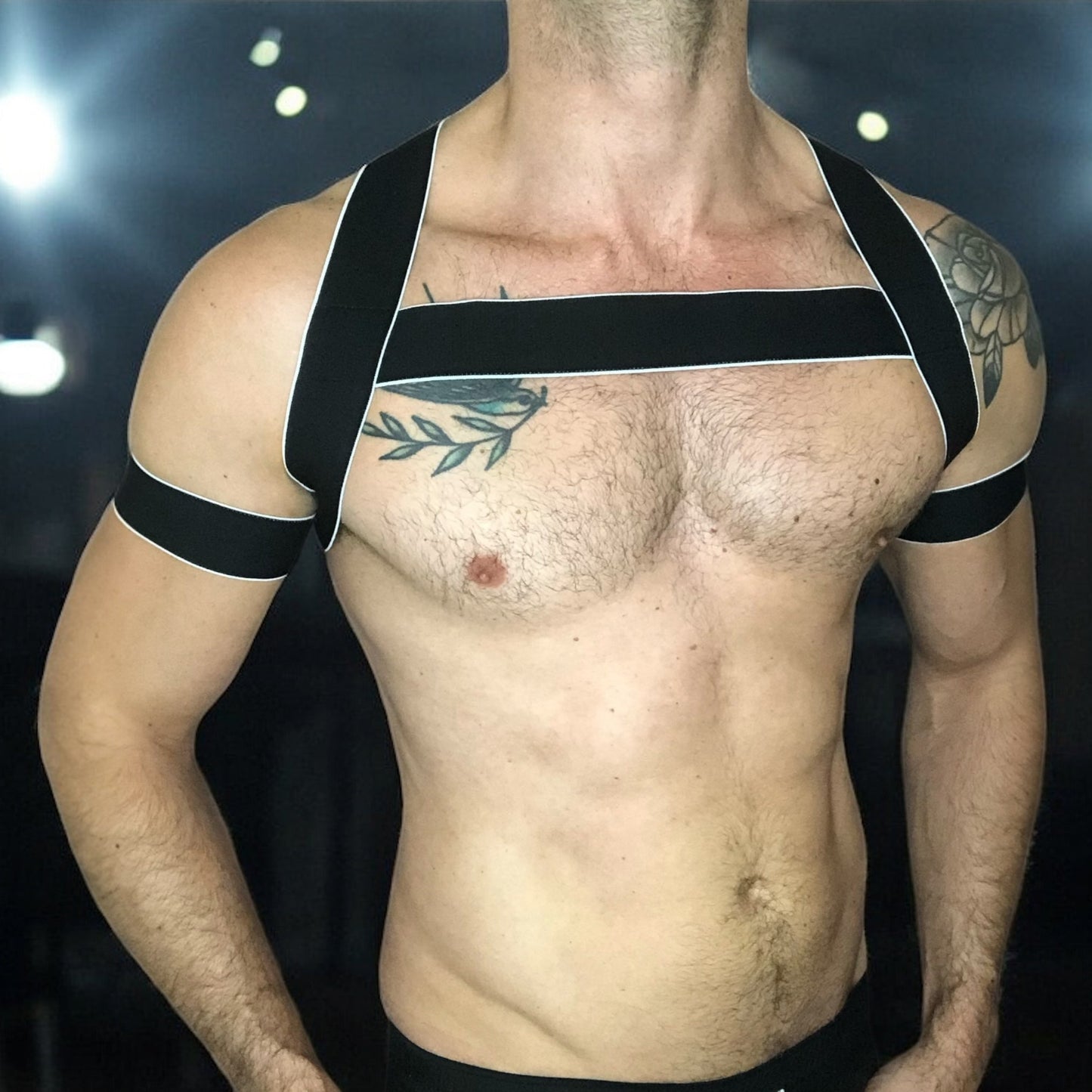 Man wearing a handmade black elastic chest harness with white trim and matching bicep bands by Cheeky Boy Harness, modeled in a dark clubwear setting.