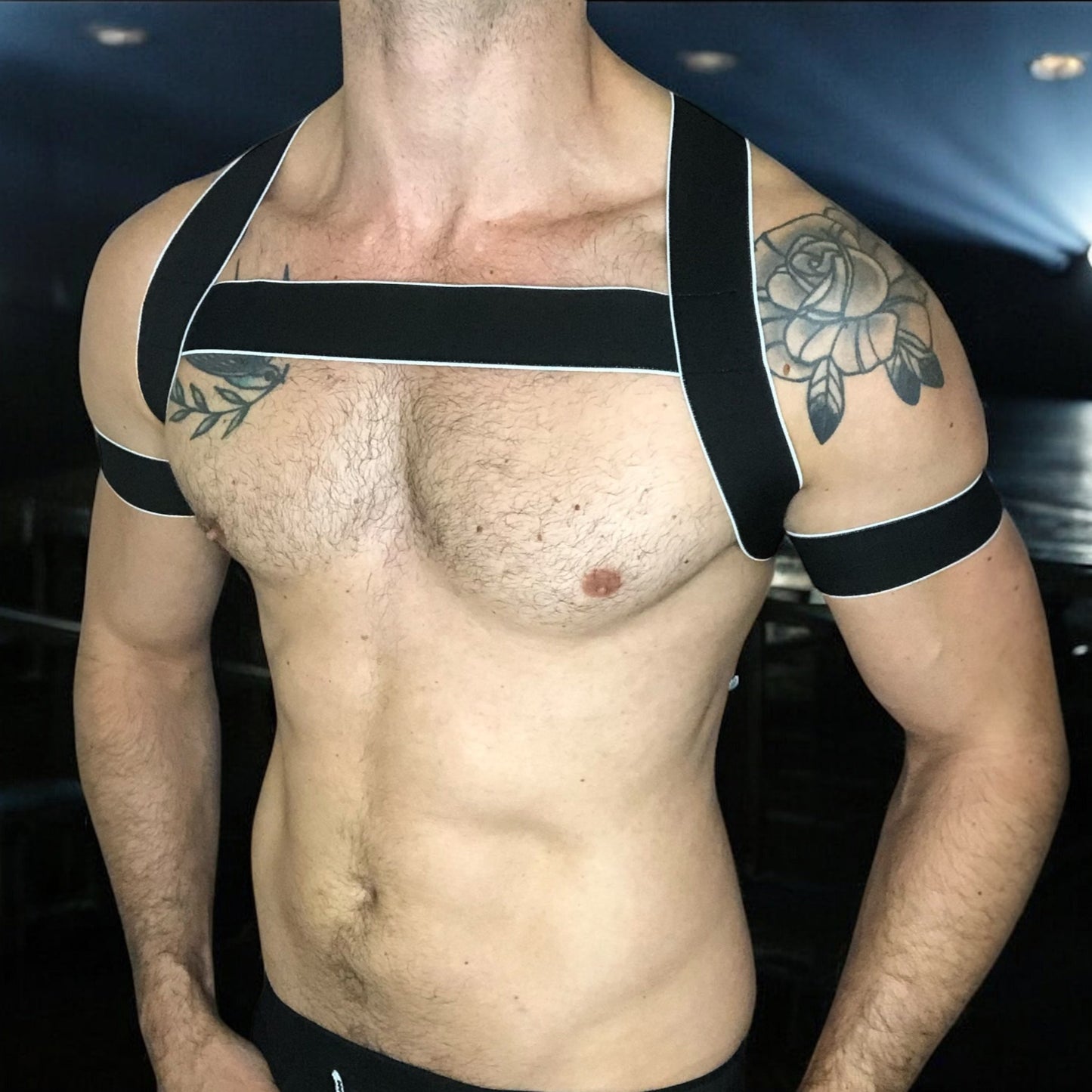 Man wearing a handmade black elastic chest harness with white trim and matching bicep bands by Cheeky Boy Harness, modeled in a dark clubwear setting.