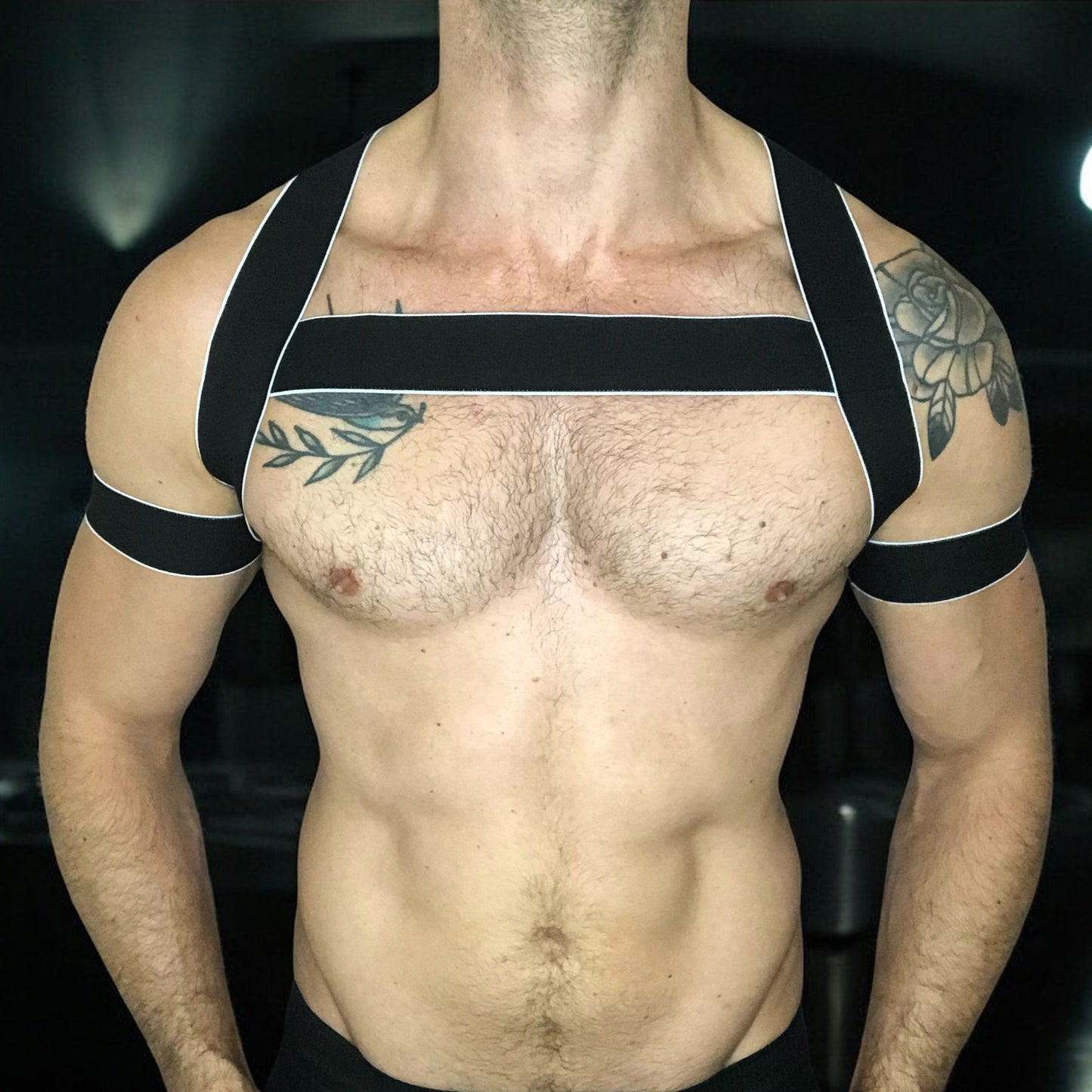 Man wearing a handmade black elastic chest harness with white trim and matching bicep bands by Cheeky Boy Harness, modeled in a dark clubwear setting.