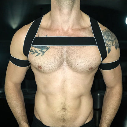 Man wearing a handmade black elastic chest harness with white trim and matching bicep bands by Cheeky Boy Harness, modeled in a dark clubwear setting.
