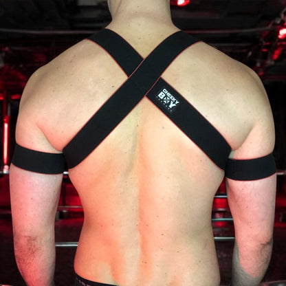 Toned model Wearing Made-to-Measure Black and Red Elastic Chest Harness with Metal Rings by Cheeky Boy Harness, Styled for Fetish Clubwear in Red-Lit Venue