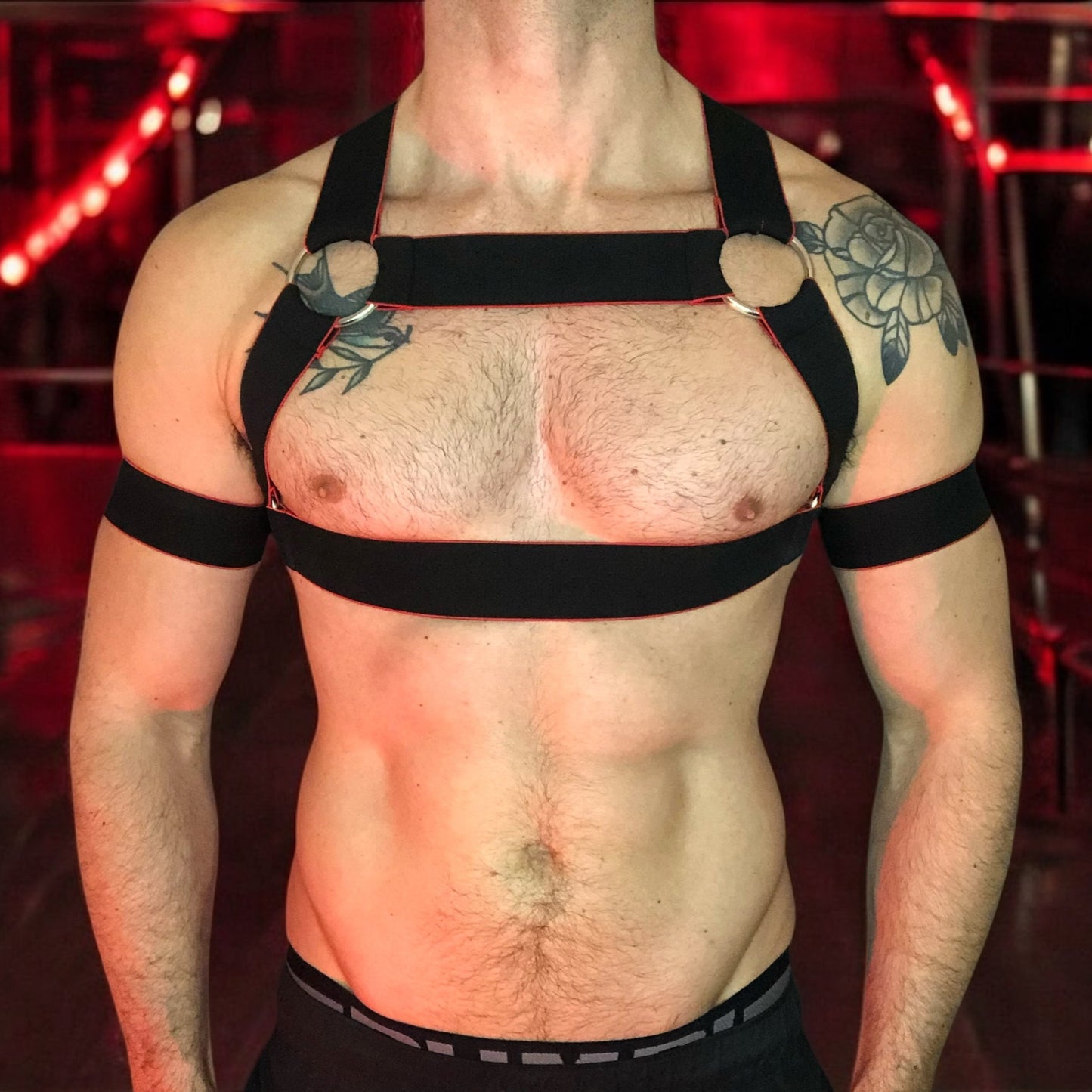Toned model Wearing Made-to-Measure Black and Red Elastic Chest Harness with Metal Rings by Cheeky Boy Harness, Styled for Fetish Clubwear in Red-Lit Venue