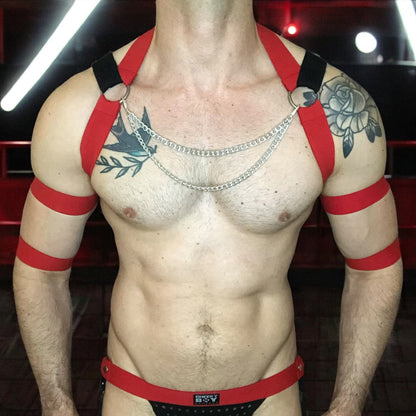 Model wearing a custom-fit black and red elastic chest harness with silver body chain detail, 4 matching bicep bands, and mesh jockstrap, photographed in a dark nightclub setting.