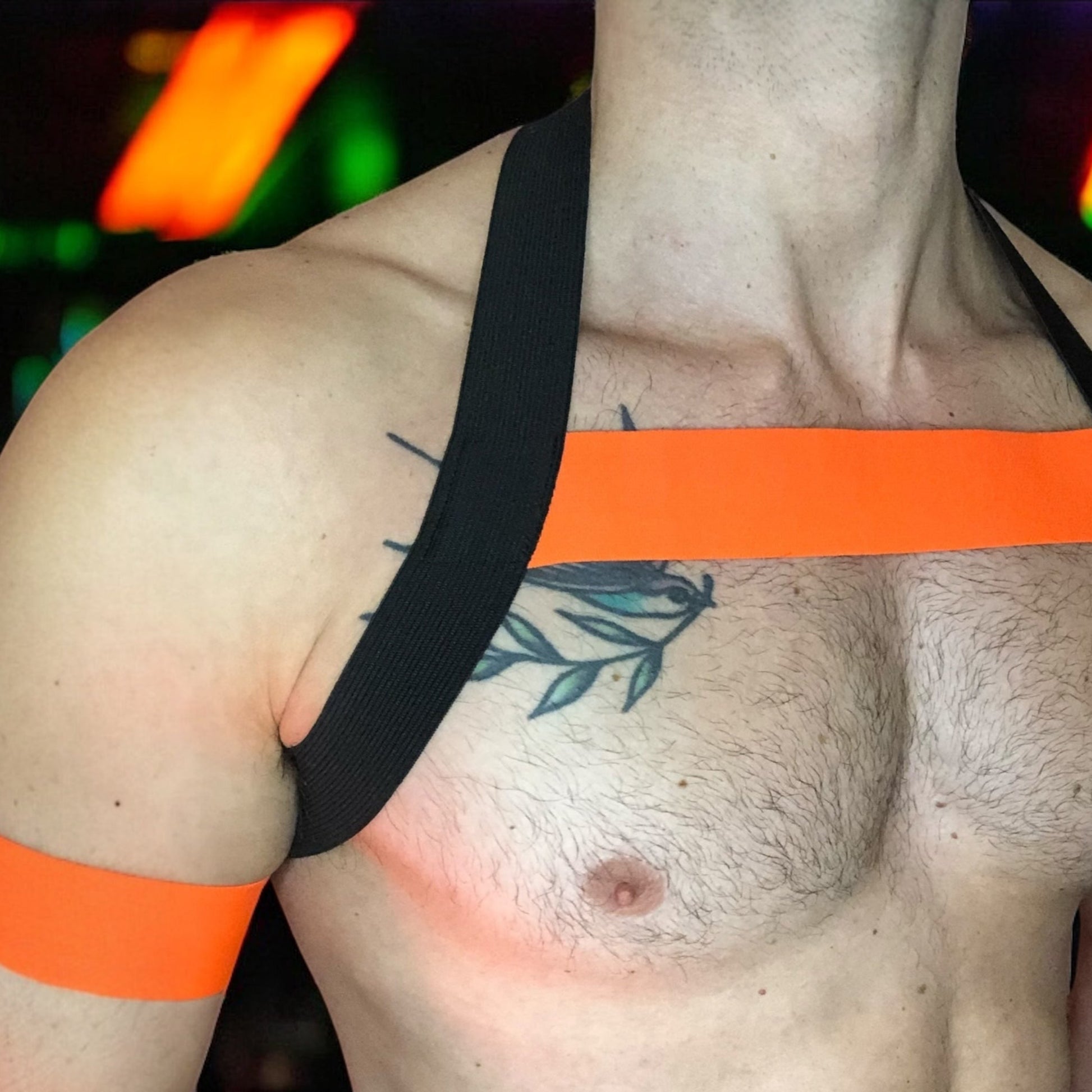 A man wearing a handmade, made-to-measure black and neon pink elastic chest harness with matching jockstrap and bicep bands by Cheeky Boy Harness, shown in a dark club setting with glowing pink UV lights