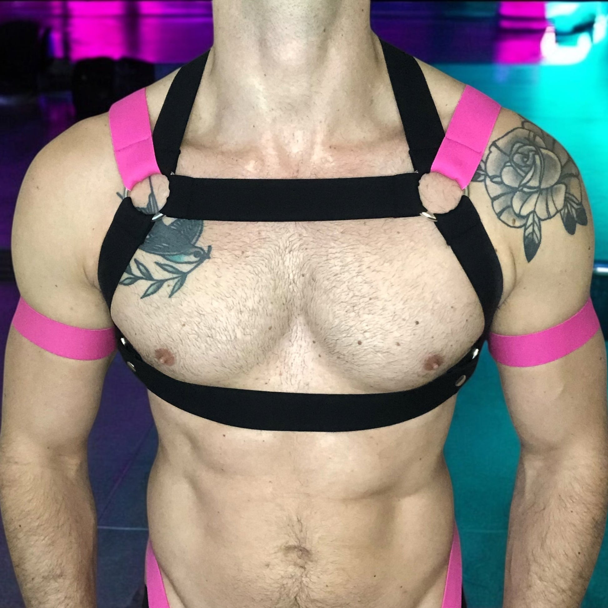 Model wearing a Cheeky Boy Harness custom-fit mens black and hot pink elastic chest and body harness, paired with matching bicep bands, leg straps, and a black mesh jockstrap. The outfit is styled for clubwear or fetish events.