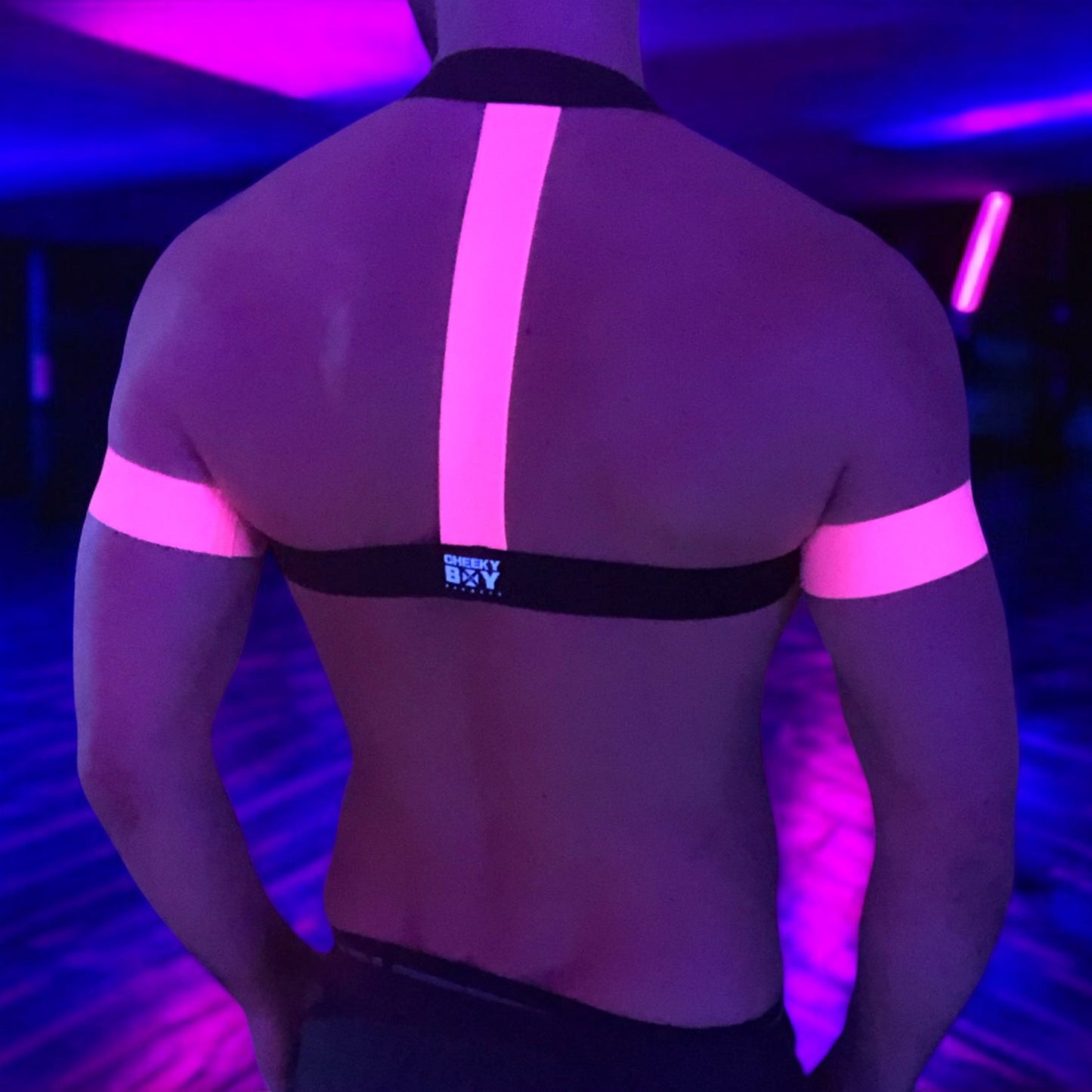A man wearing a handmade, made-to-measure black and neon pink elastic chest harness with matching jockstrap and bicep bands by Cheeky Boy Harness, shown in a dark club setting with glowing pink UV lights