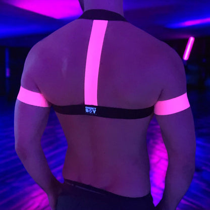 A man wearing a handmade, made-to-measure black and neon pink elastic chest harness with matching jockstrap and bicep bands by Cheeky Boy Harness, shown in a dark club setting with glowing pink UV lights