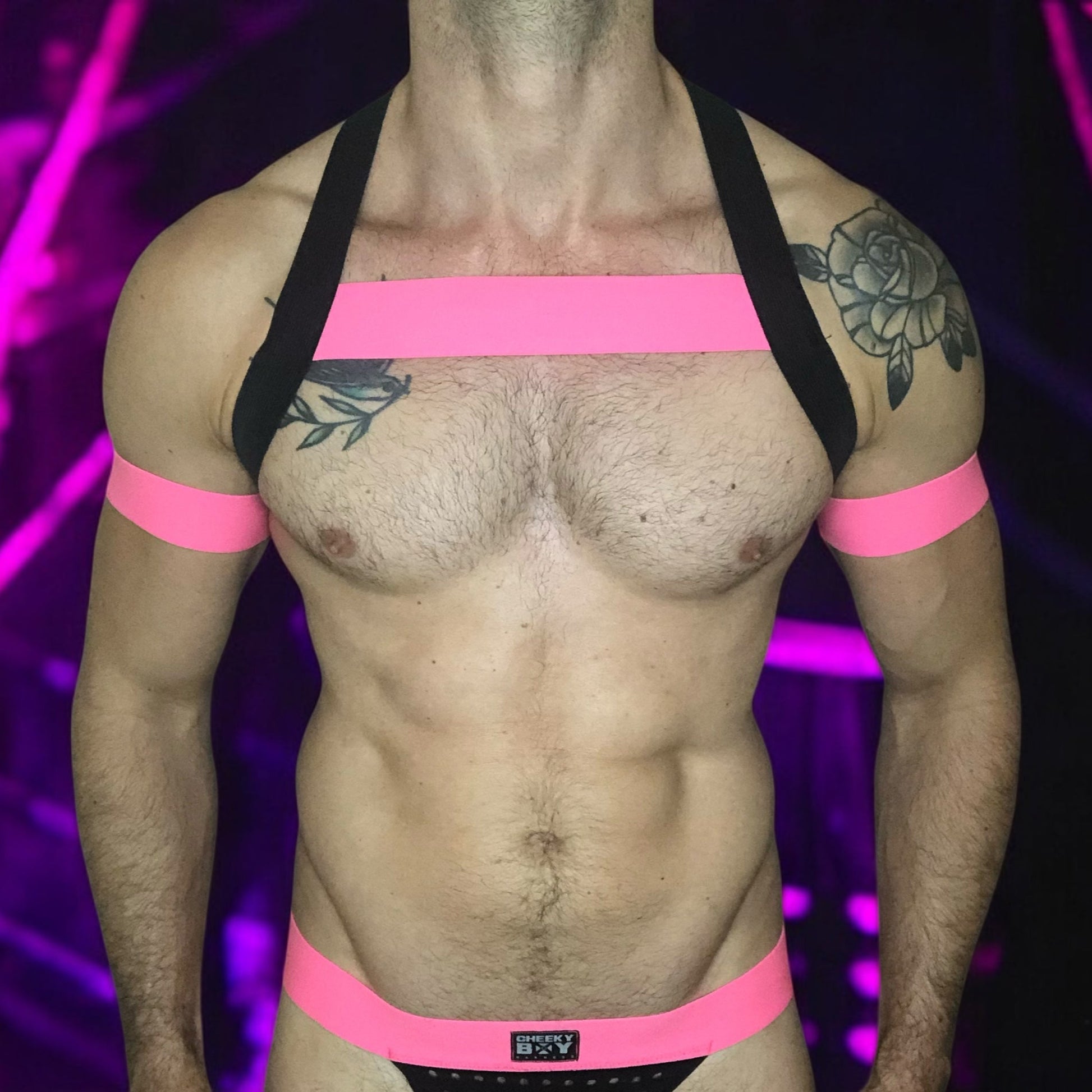 A man wearing a handmade, made-to-measure black and neon pink elastic chest harness with matching jockstrap and bicep bands by Cheeky Boy Harness, shown in a dark club setting with glowing pink UV lights