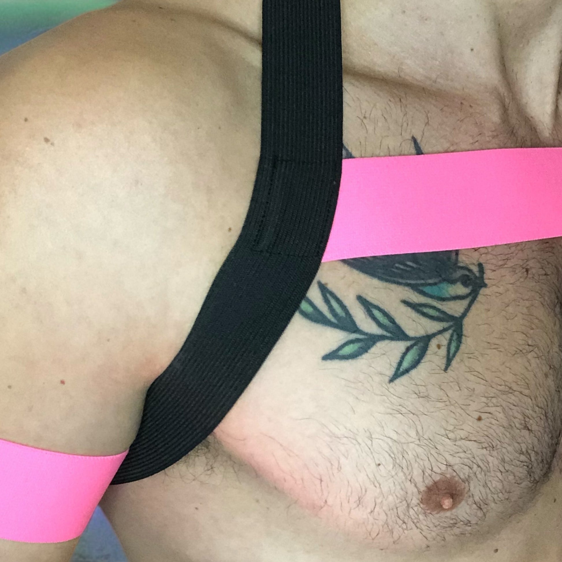 A man wearing a handmade, made-to-measure black and neon pink elastic chest harness with matching jockstrap and bicep bands by Cheeky Boy Harness, shown in a dark club setting with glowing pink UV lights