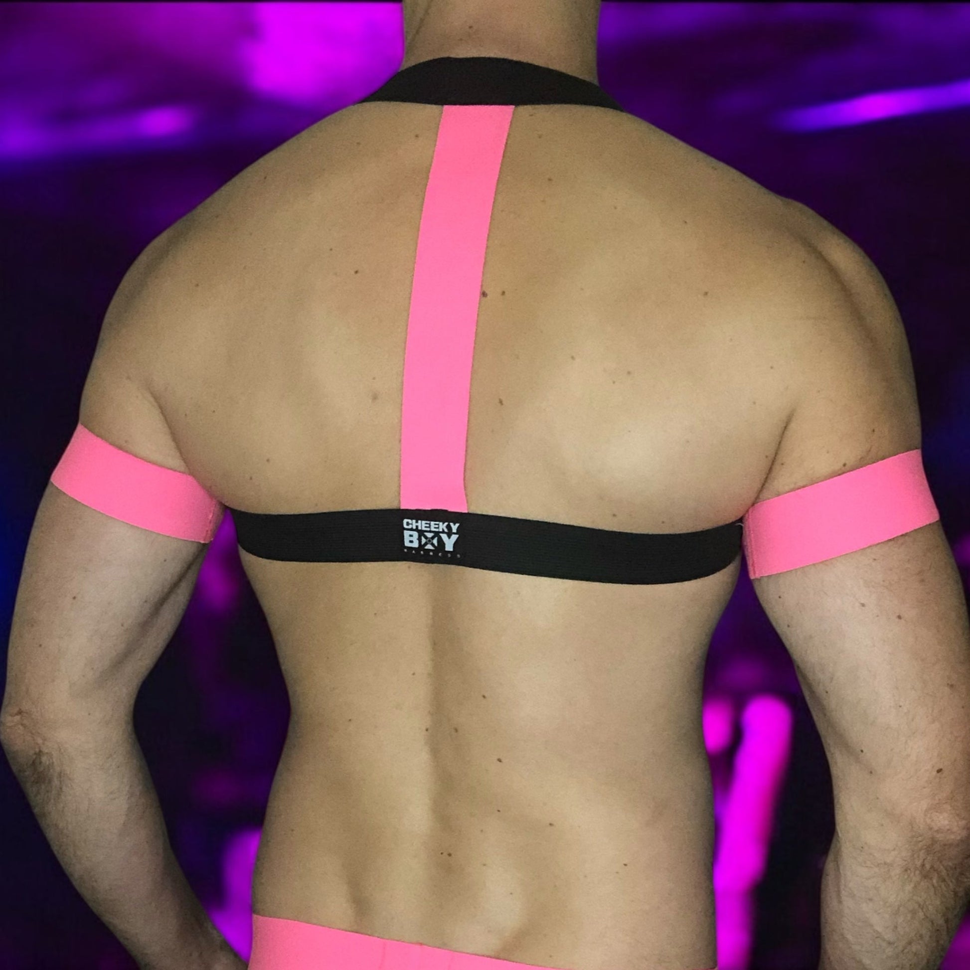 A man wearing a handmade, made-to-measure black and neon pink elastic chest harness with matching jockstrap and bicep bands by Cheeky Boy Harness, shown in a dark club setting with glowing pink UV lights