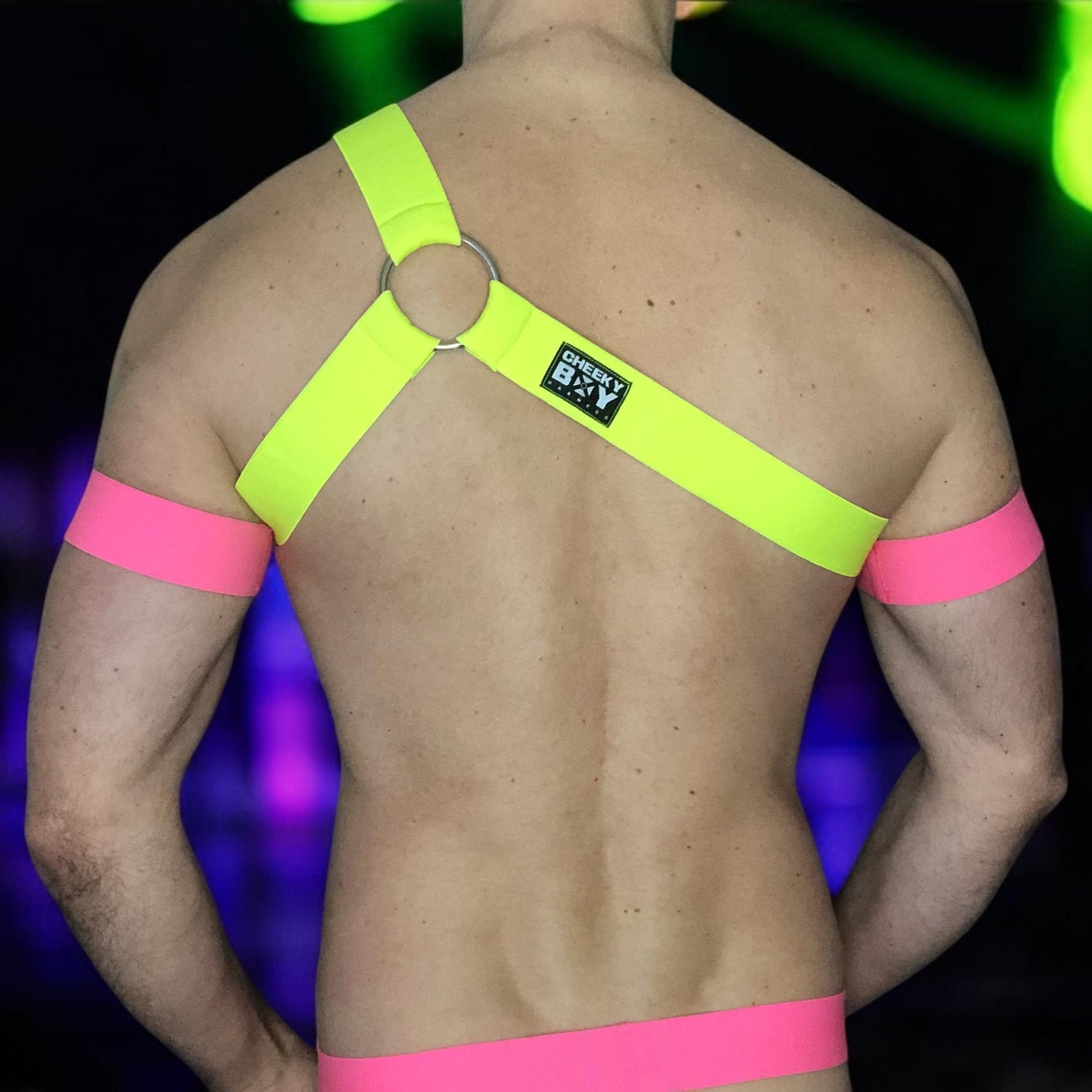 Model wearing a custom-fit Cheeky Boy Harness set featuring a neon yellow one-shoulder chest harness with a silver ring, paired with pink elastic bicep bands and waistband, and a black mesh jockstrap. Shot under UV lighting in a club setting.