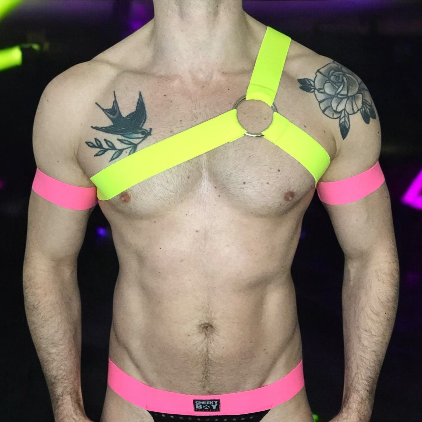 Model wearing a custom-fit Cheeky Boy Harness set featuring a neon yellow one-shoulder chest harness with a silver ring, paired with pink elastic bicep bands and waistband, and a black mesh jockstrap. Shot under UV lighting in a club setting.