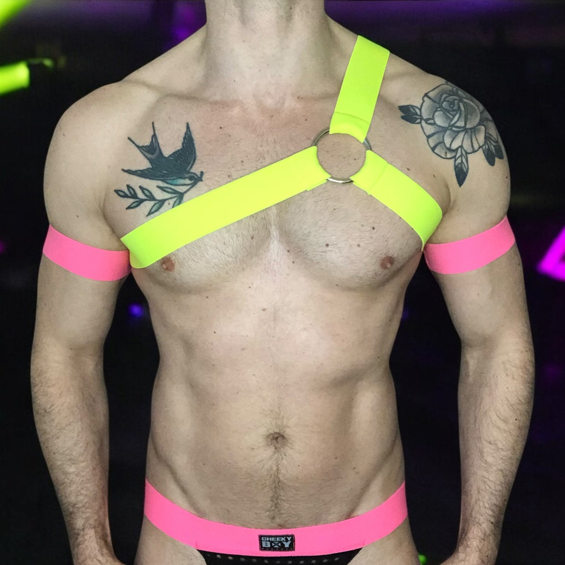 Model wearing a custom-fit Cheeky Boy Harness set featuring a neon yellow one-shoulder chest harness with a silver ring, paired with pink elastic bicep bands and waistband, and a black mesh jockstrap. Shot under UV lighting in a club setting.