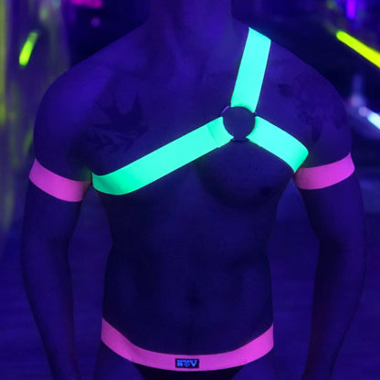 Model wearing a custom-fit Cheeky Boy Harness set featuring a neon yellow one-shoulder chest harness with a silver ring, paired with pink elastic bicep bands and waistband, and a black mesh jockstrap. Shot under UV lighting in a club setting.
