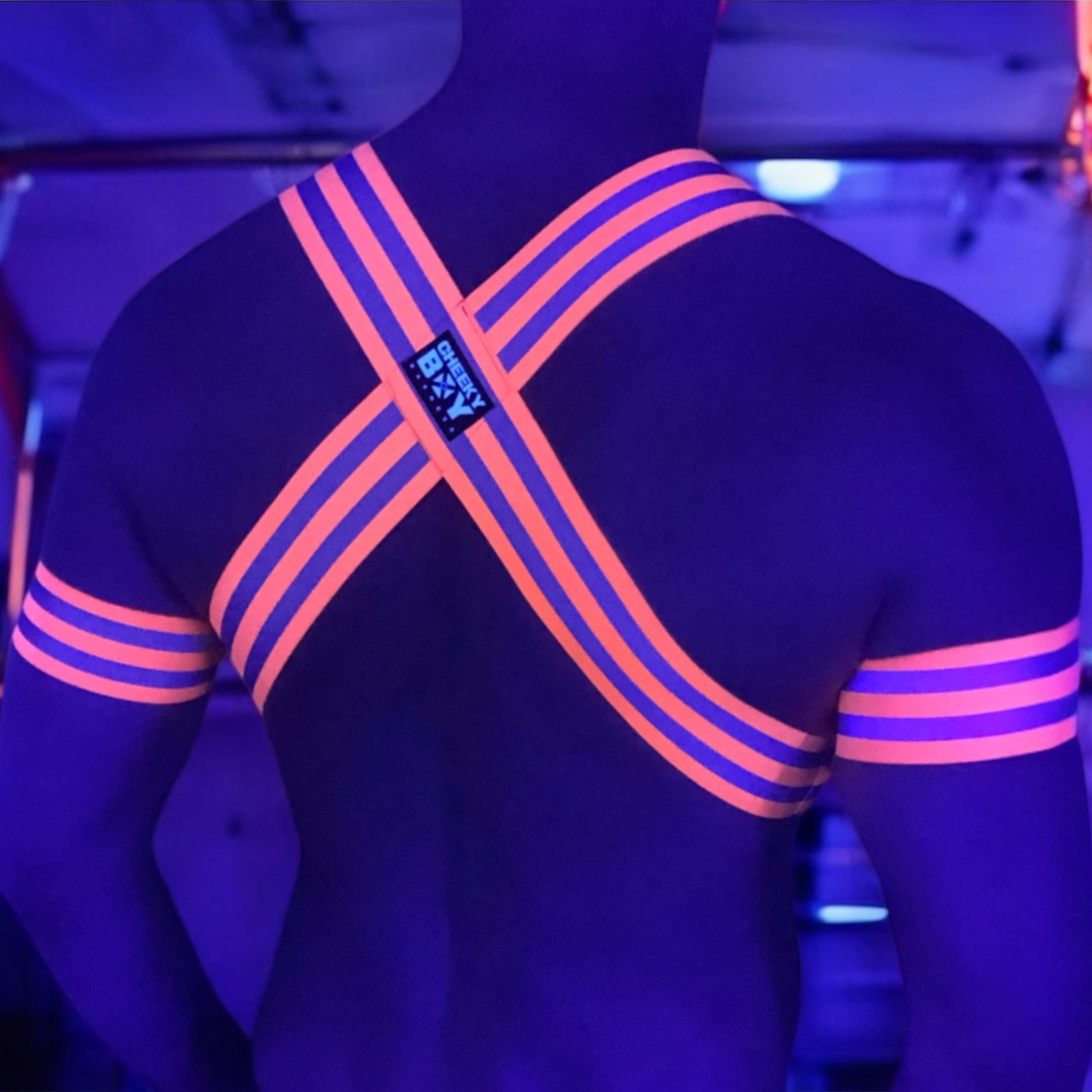 Made-to-measure men’s elastic chest harness in high-visibility neon orange with reflective detailing, glowing under blacklight. The design features double shoulder and chest straps joined by circular O-rings, with optional matching jockstrap and thigh bands—ideal for clubwear, festivals, or fetish fashion.