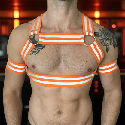 Made-to-measure men’s elastic chest harness in high-visibility neon orange with reflective detailing, glowing under blacklight. The design features double shoulder and chest straps joined by circular O-rings, with optional matching jockstrap and thigh bands—ideal for clubwear, festivals, or fetish fashion.